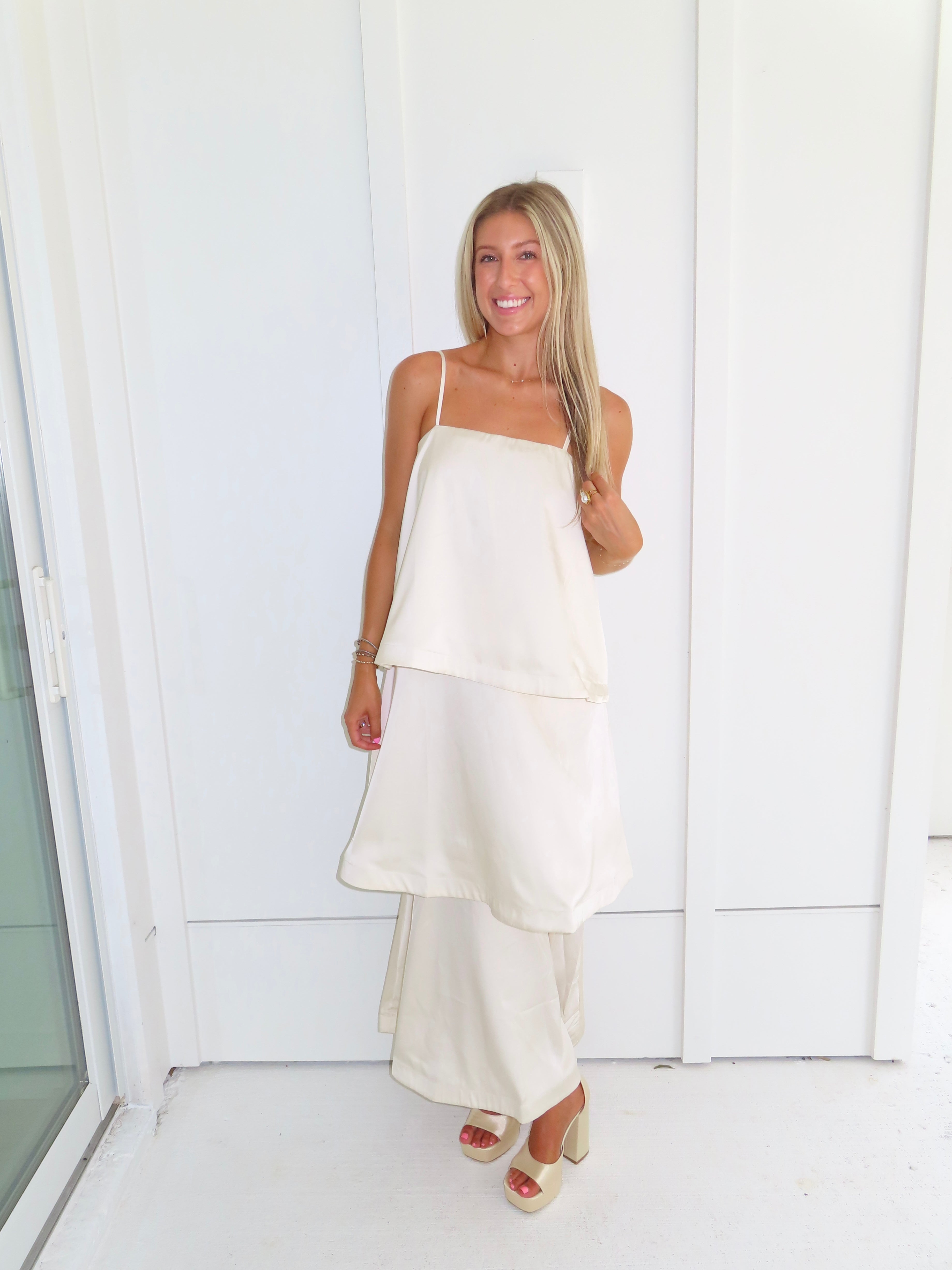 A woman modeling a cream-colored tiered  layered maxi dress and gold heels standing against a white paneled wall.