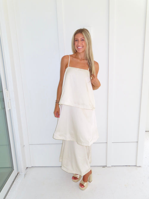 A woman modeling a cream-colored tiered  layered maxi dress and gold heels standing against a white paneled wall.