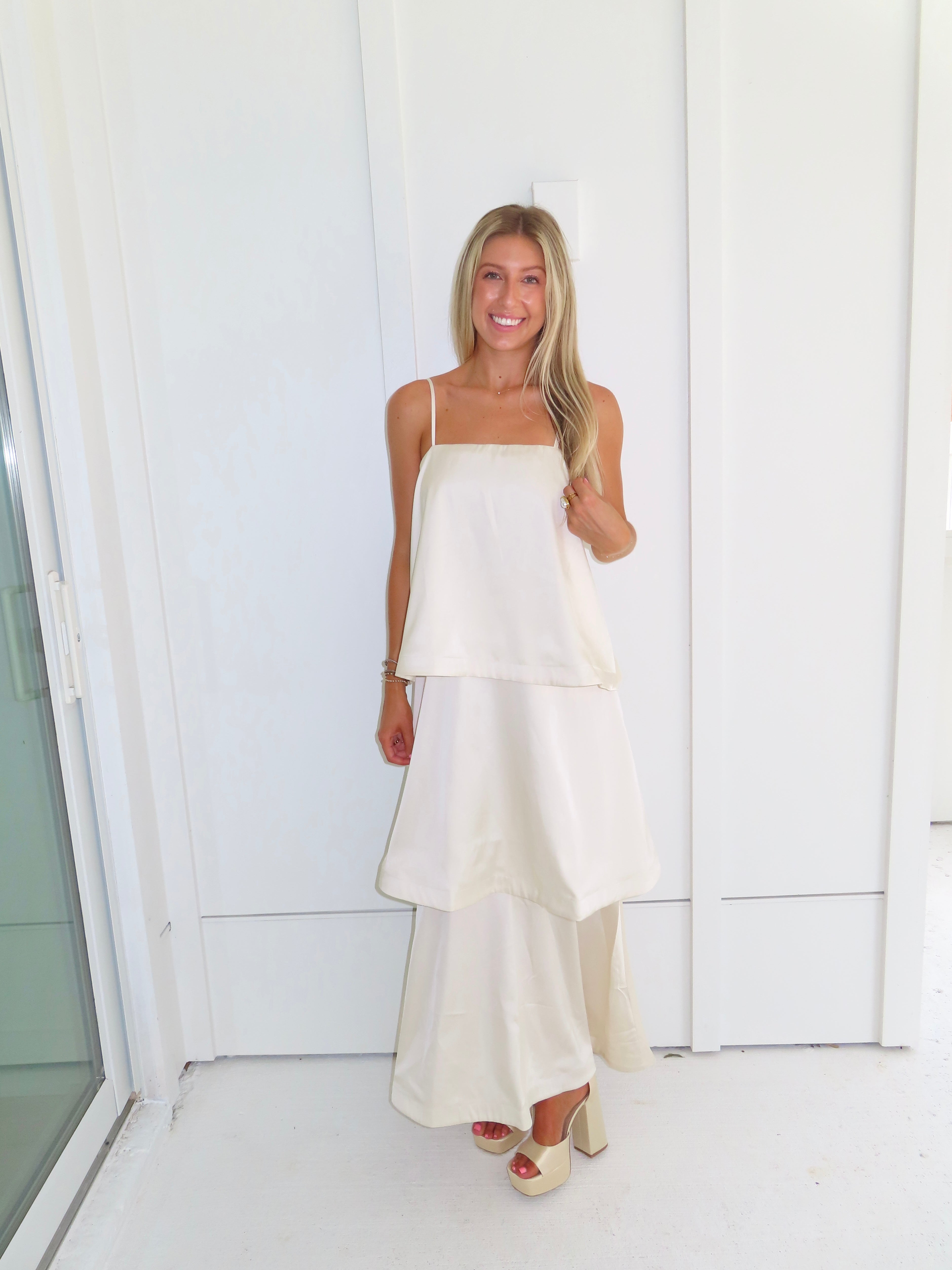 A woman modeling a cream-colored tiered  layered maxi dress and gold heels standing against a white paneled wall.