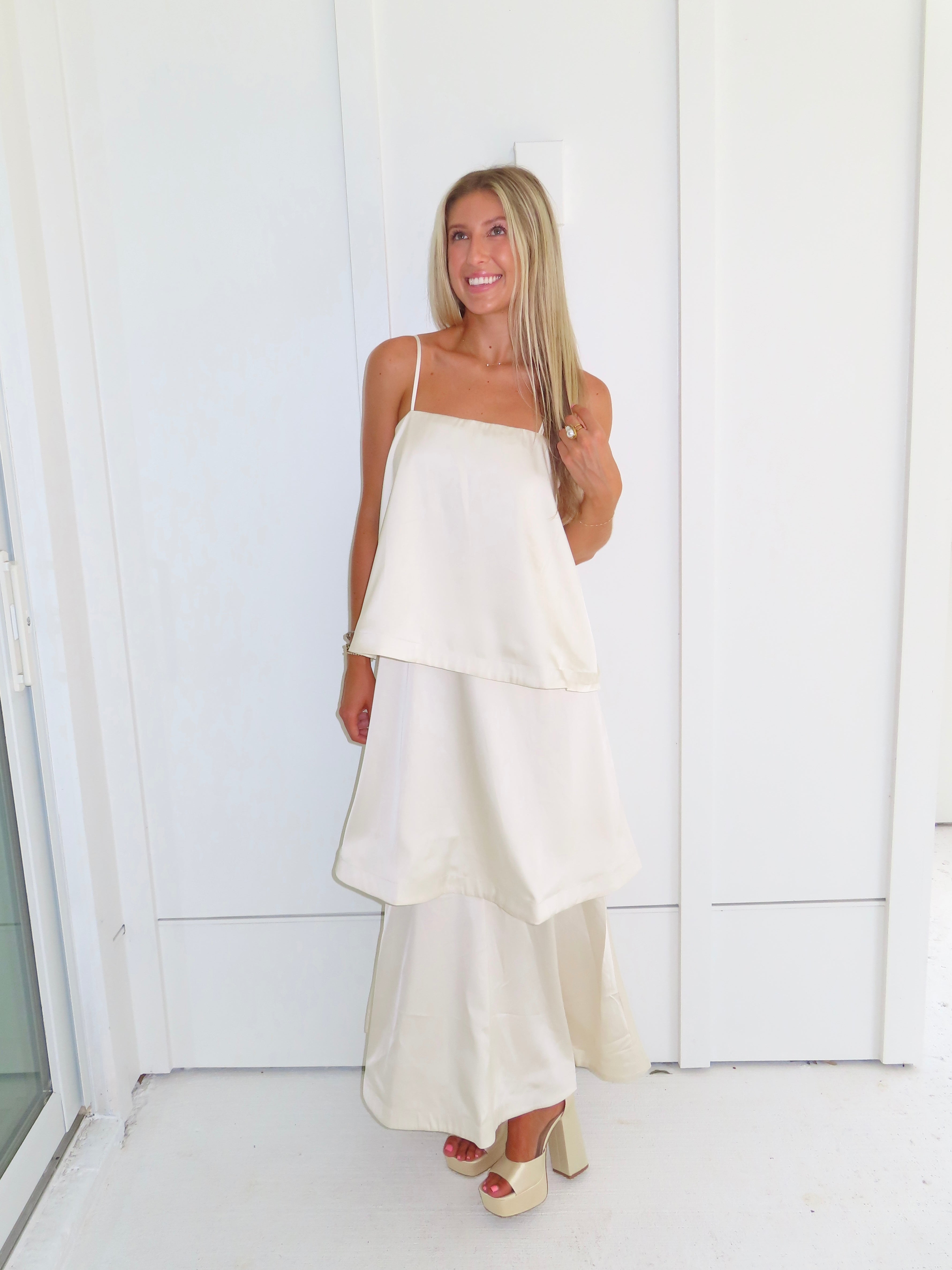 A woman modeling a cream-colored tiered  layered maxi dress and gold heels standing against a white paneled wall.