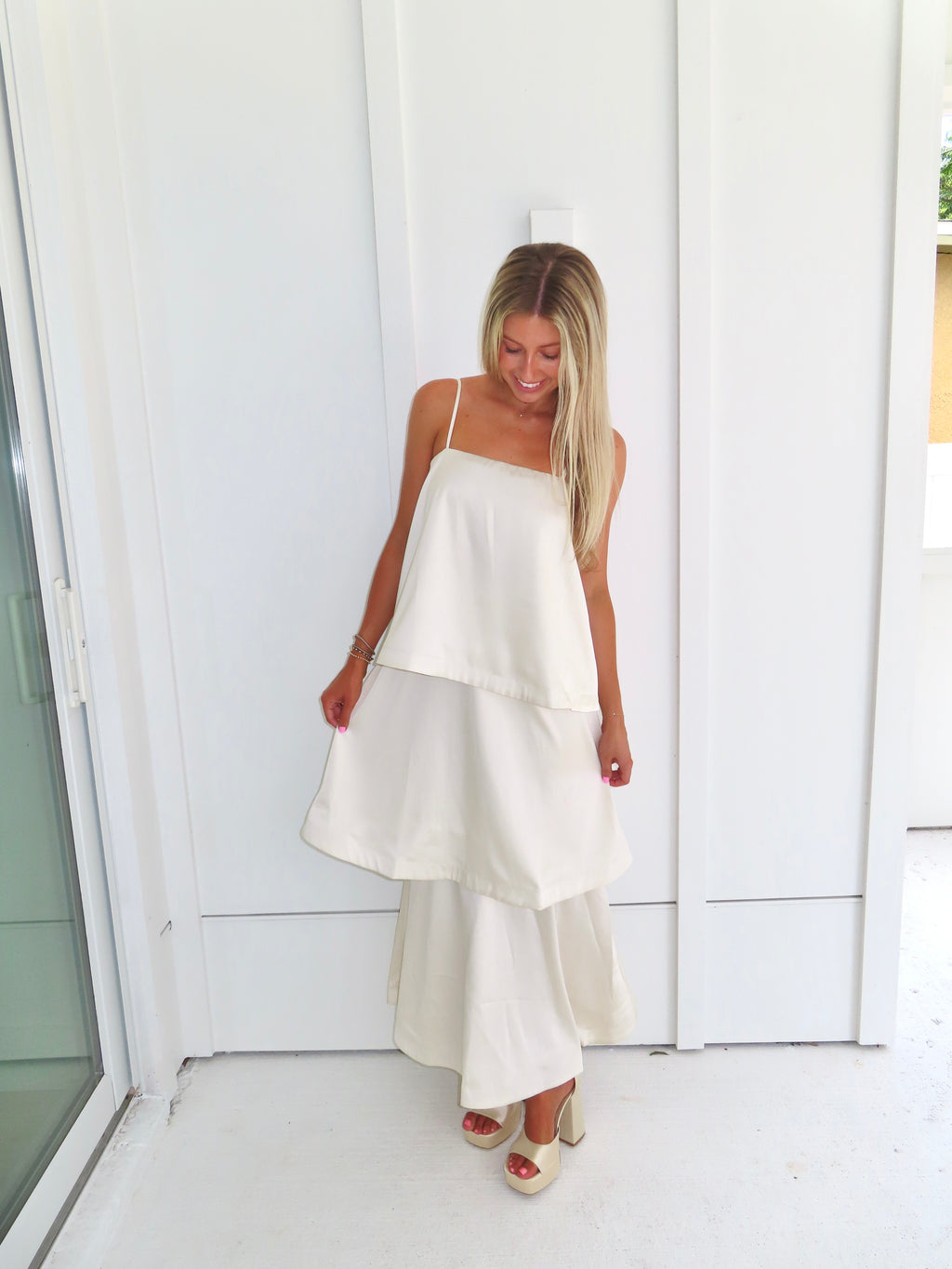A woman modeling a cream-colored tiered  layered maxi dress and gold heels standing against a white paneled wall.