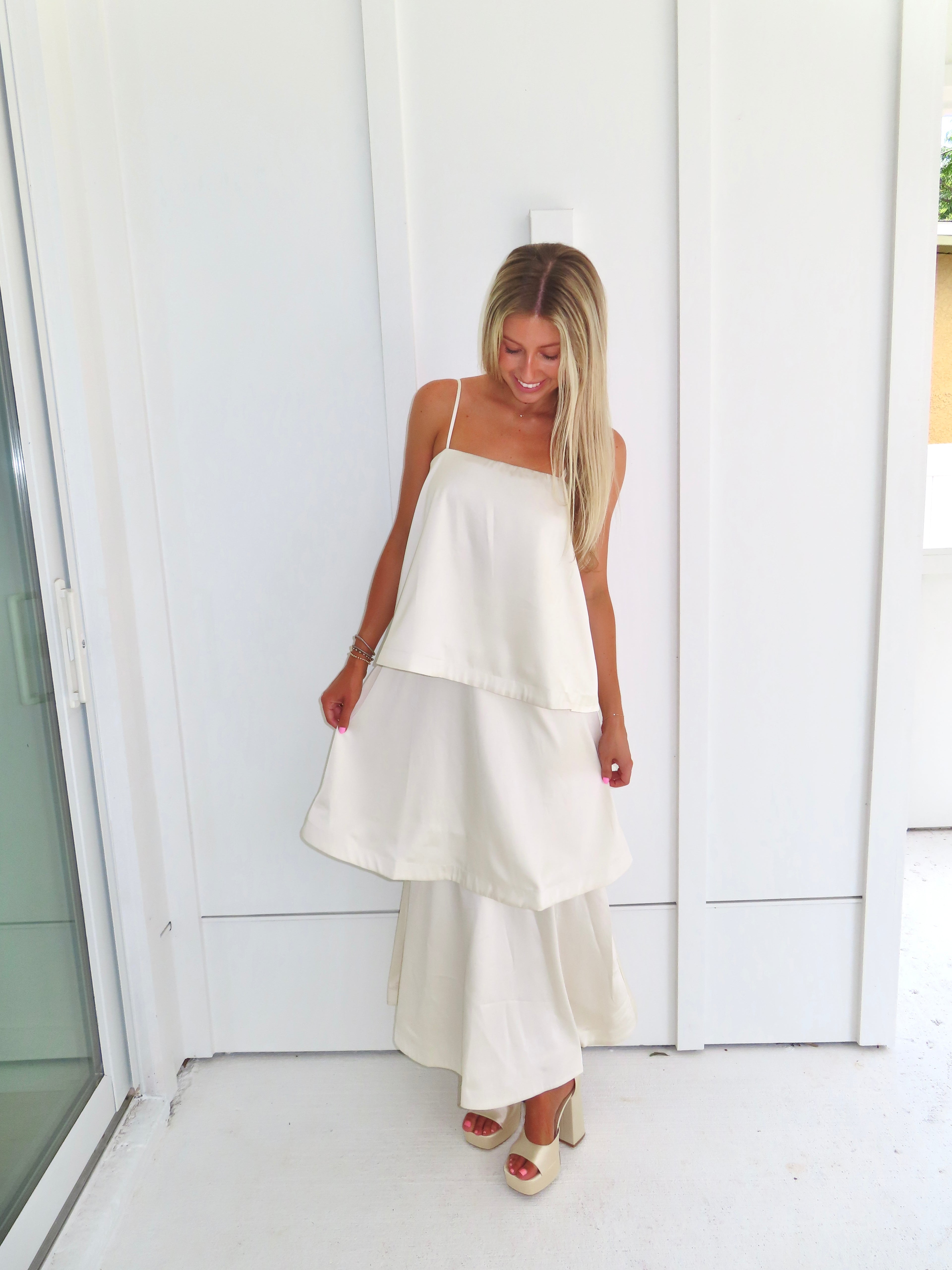 A woman modeling a cream-colored tiered  layered maxi dress and gold heels standing against a white paneled wall.