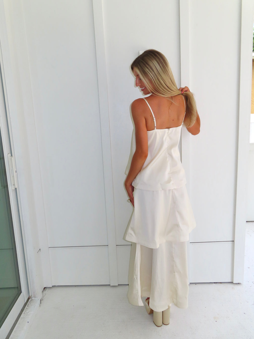 A woman modeling a cream-colored tiered  layered maxi dress and gold heels standing against a white paneled wall.