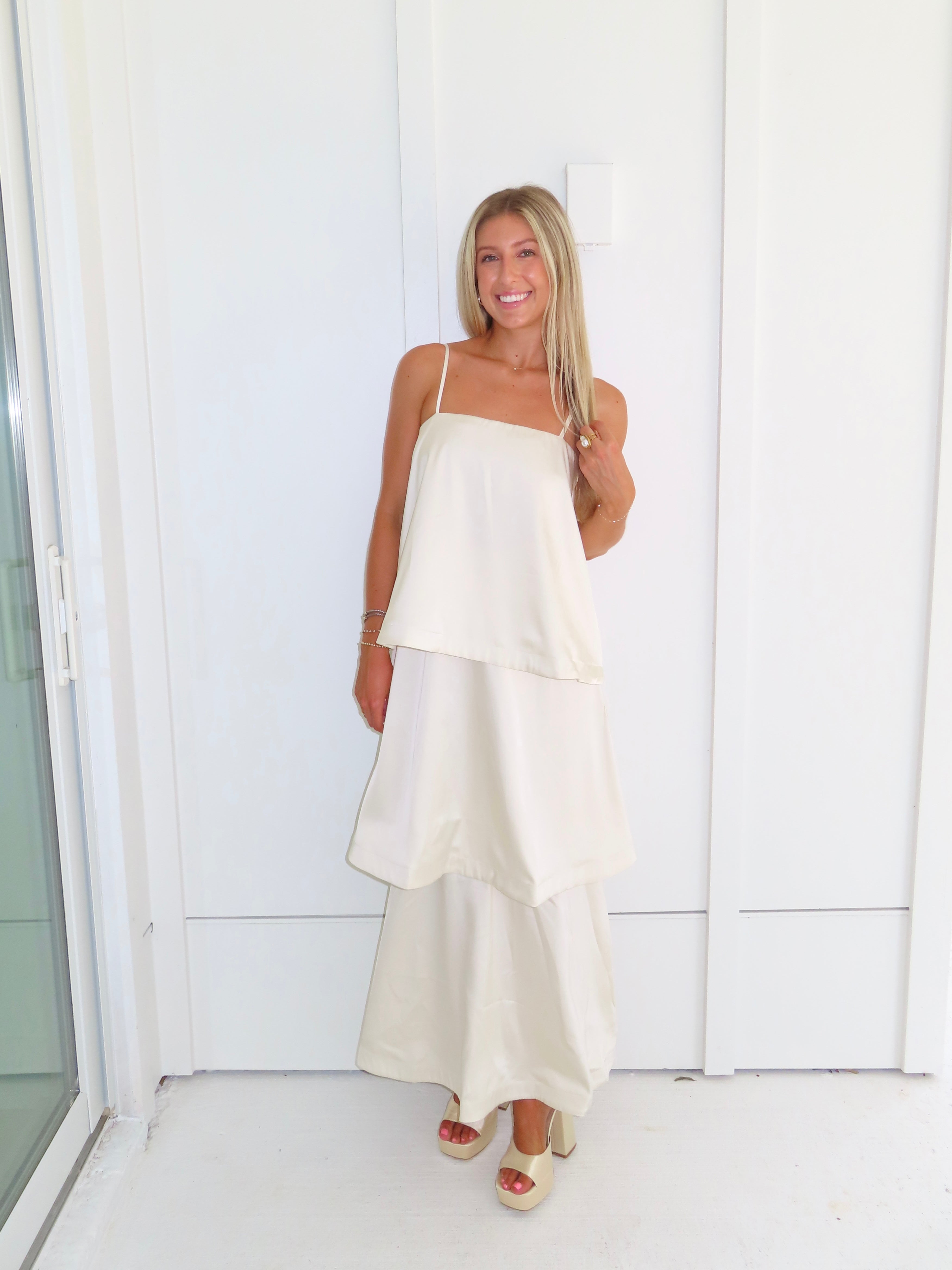 A woman modeling a cream-colored tiered  layered maxi dress and gold heels standing against a white paneled wall.