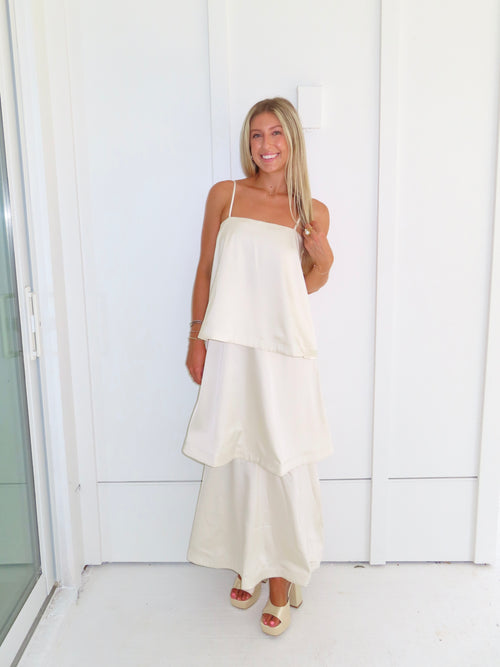 A woman modeling a cream-colored tiered  layered maxi dress and gold heels standing against a white paneled wall.
