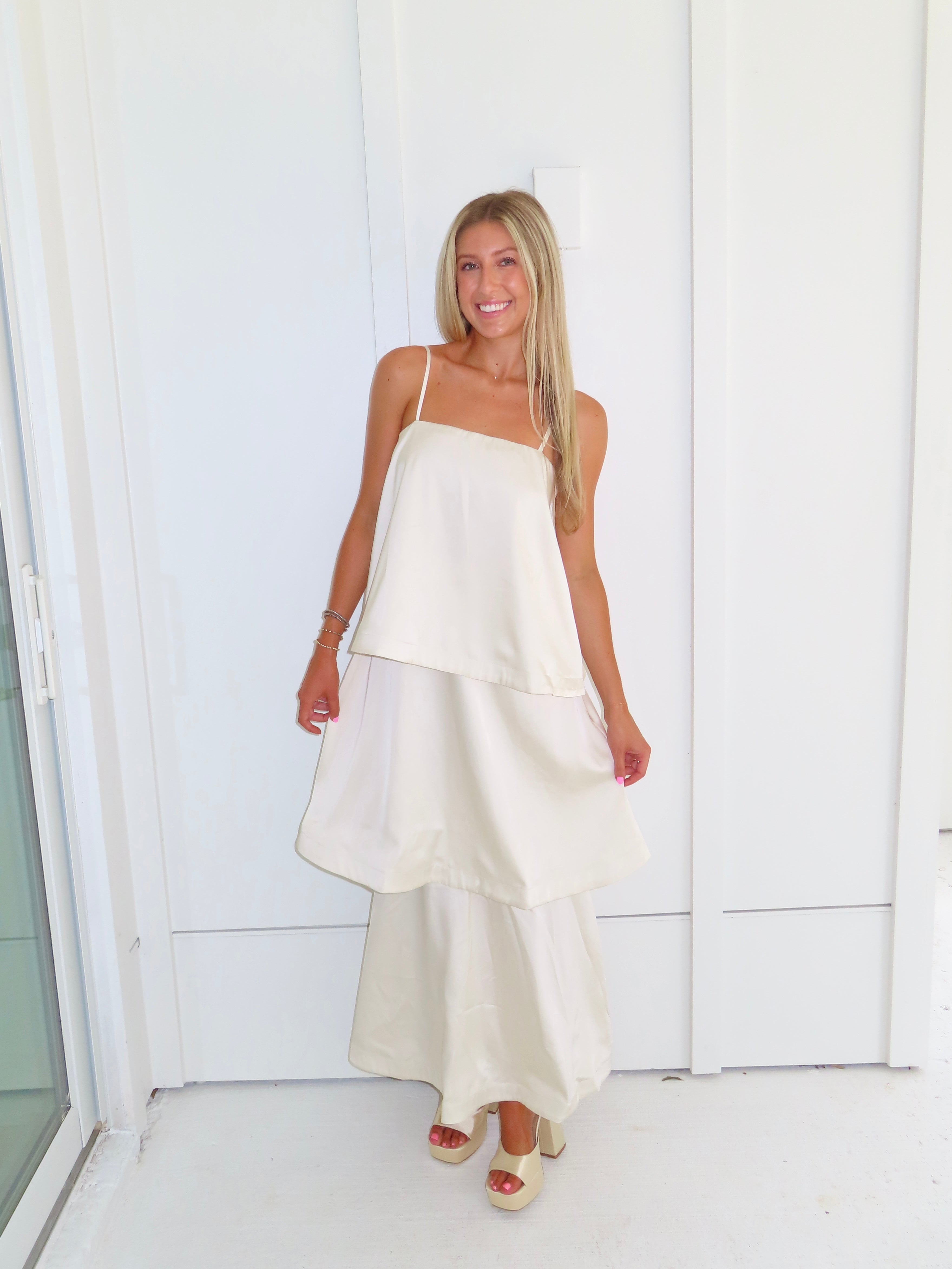 A woman modeling a cream-colored tiered  layered maxi dress and gold heels standing against a white paneled wall.