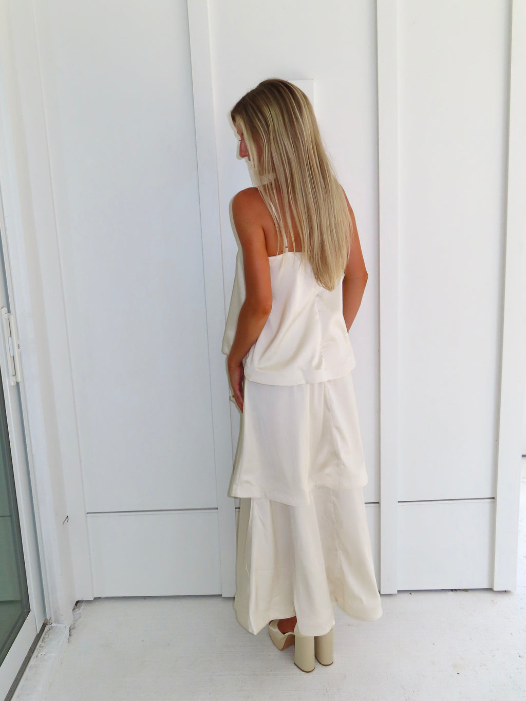 A woman modeling a cream-colored tiered  layered maxi dress and gold heels standing against a white paneled wall.