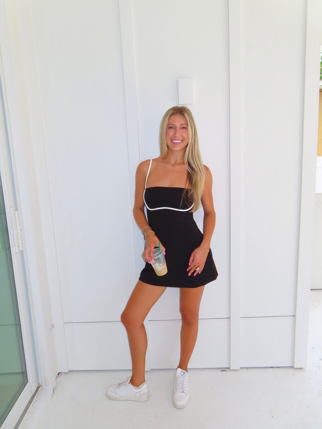 A woman posing in a black athletic dress with white trim, holding a coffee and wearing white sneakers, standing against a white paneled wall.
