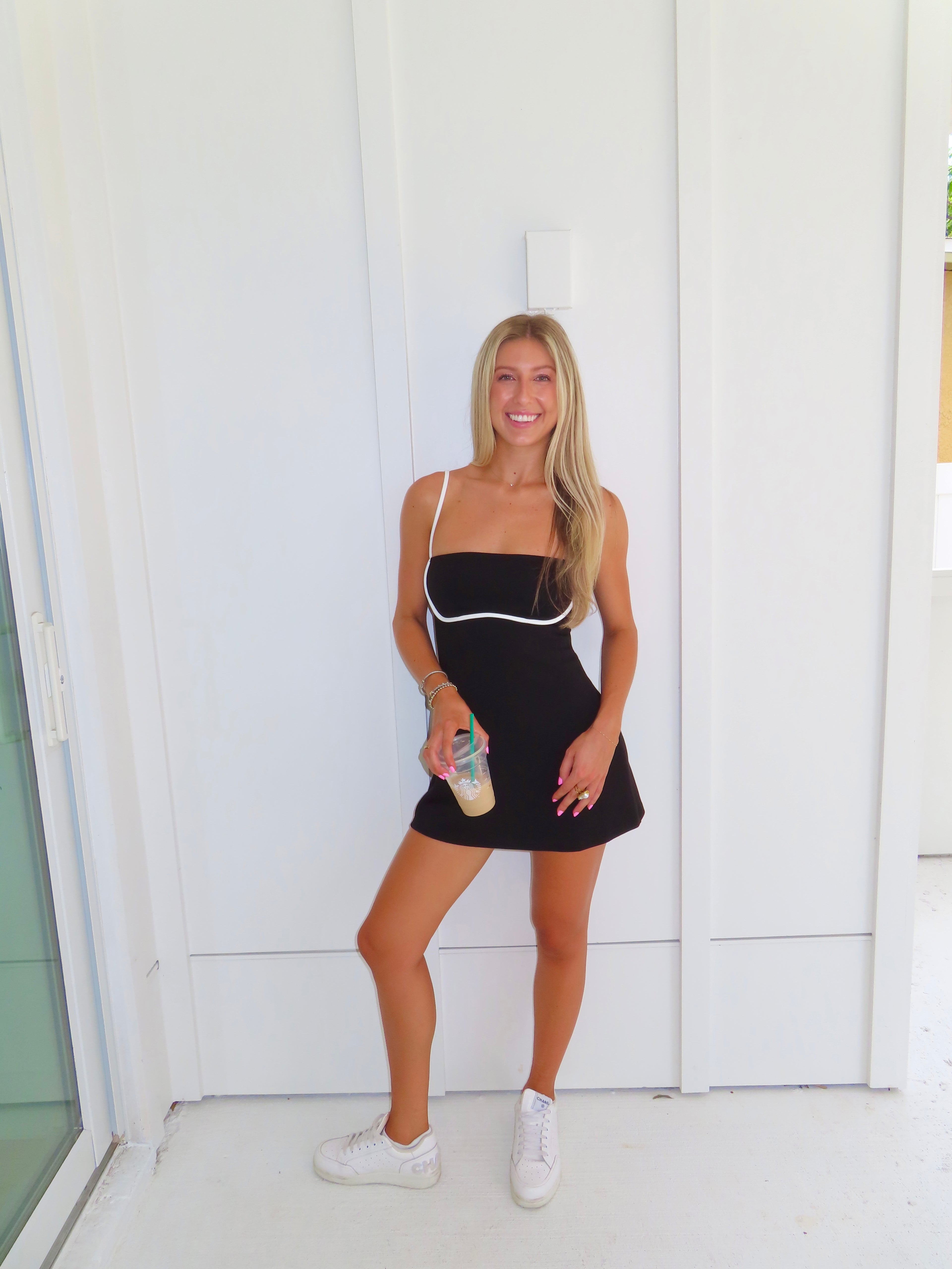 A woman posing in a black athletic dress with white trim, holding a coffee and wearing white sneakers, standing against a white paneled wall.