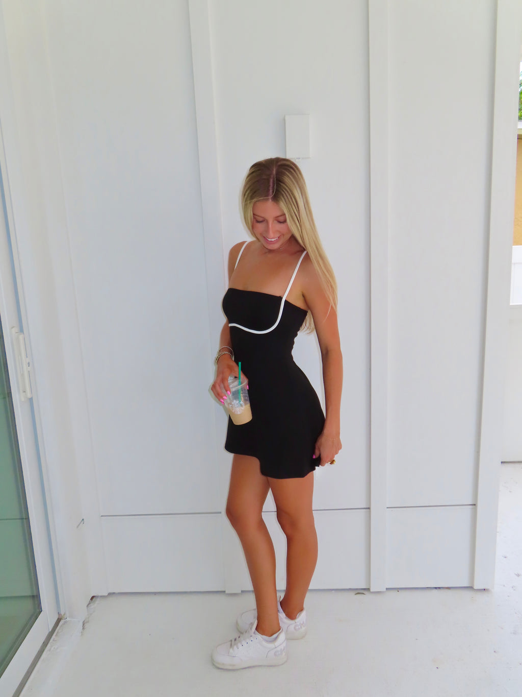 A woman posing in a black athletic dress with white trim, holding a coffee and wearing white sneakers, standing against a white paneled wall.