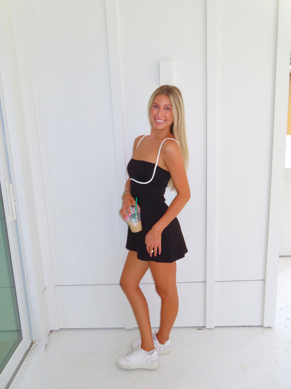 A woman posing in a black athletic dress with white trim, holding a coffee and wearing white sneakers, standing against a white paneled wall.