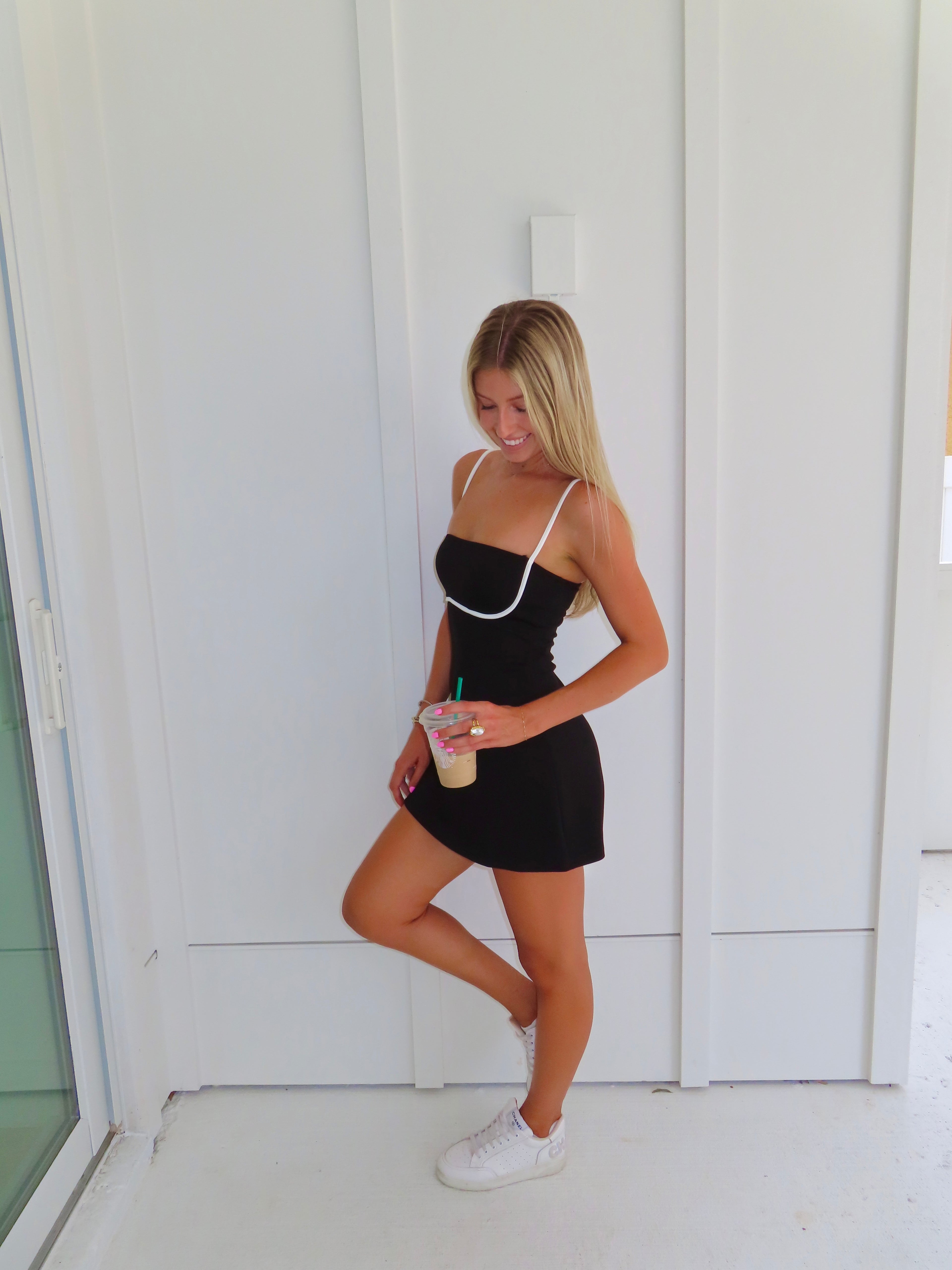 A woman posing in a black athletic dress with white trim, holding a coffee and wearing white sneakers, standing against a white paneled wall.