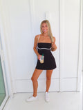 A woman posing in a black athletic dress with white trim, holding a coffee and wearing white sneakers, standing against a white paneled wall.