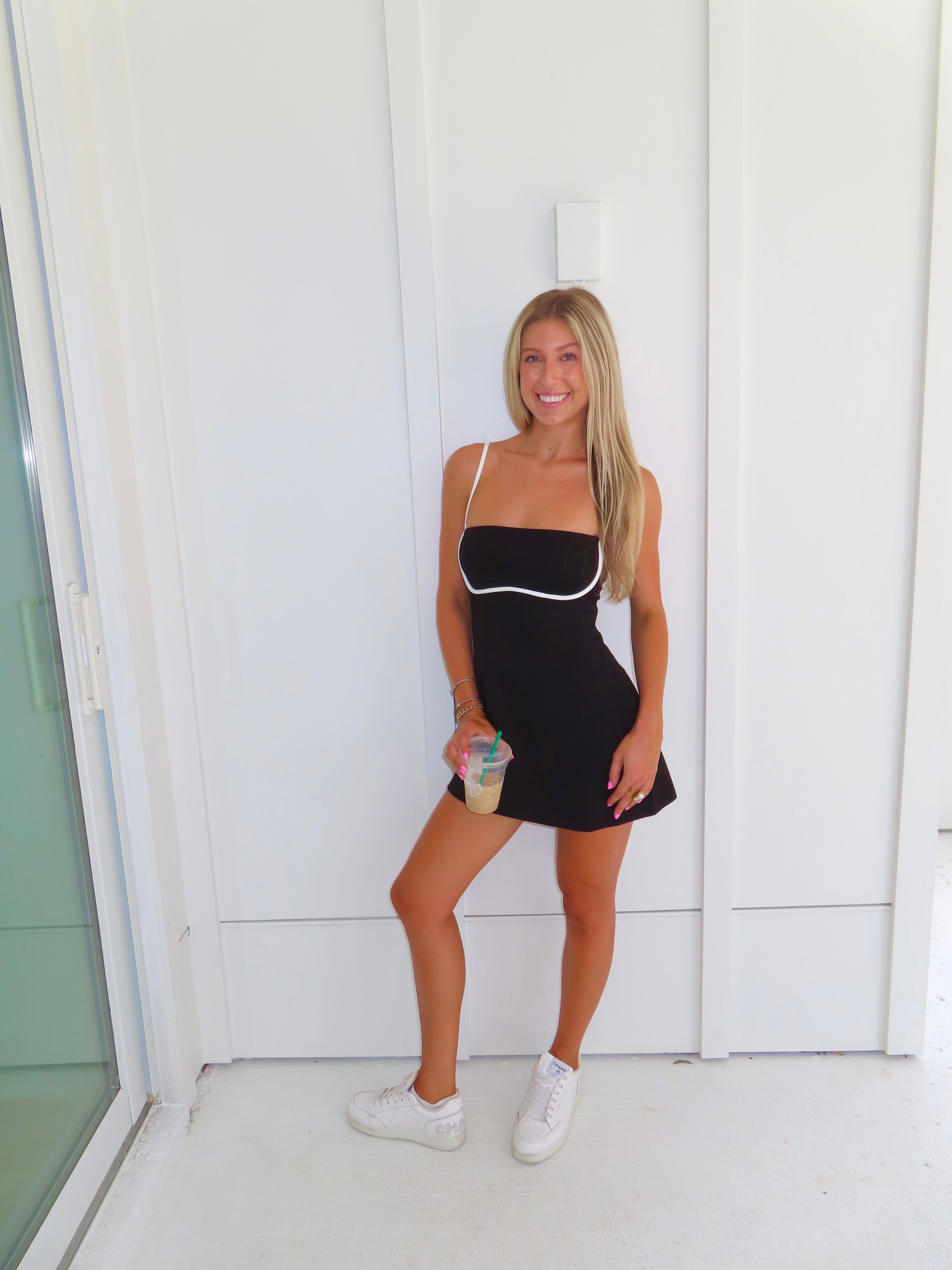 A woman posing in a black athletic dress with white trim, holding a coffee and wearing white sneakers, standing against a white paneled wall.