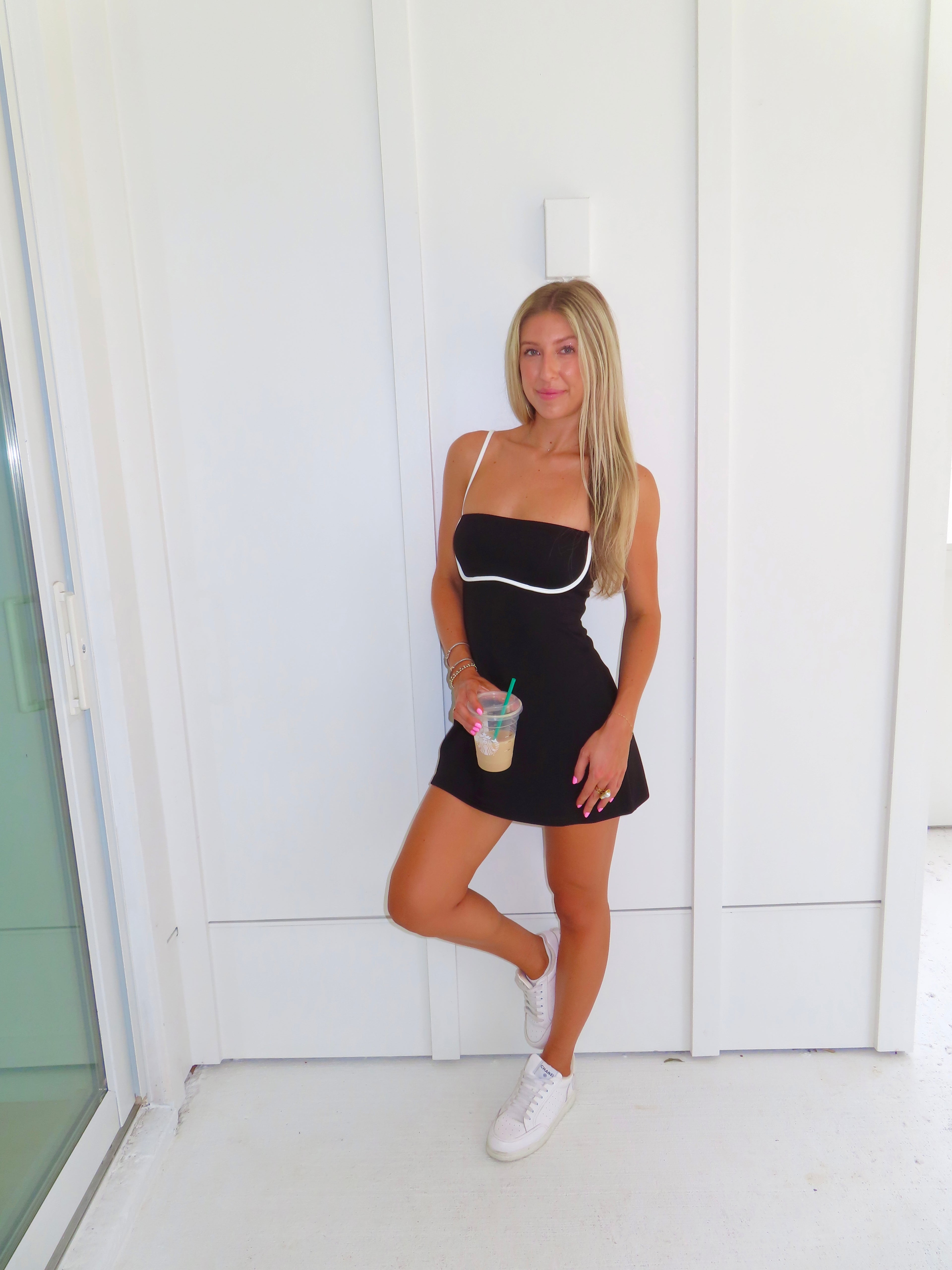 A woman posing in a black athletic dress with white trim, holding a coffee and wearing white sneakers, standing against a white paneled wall.