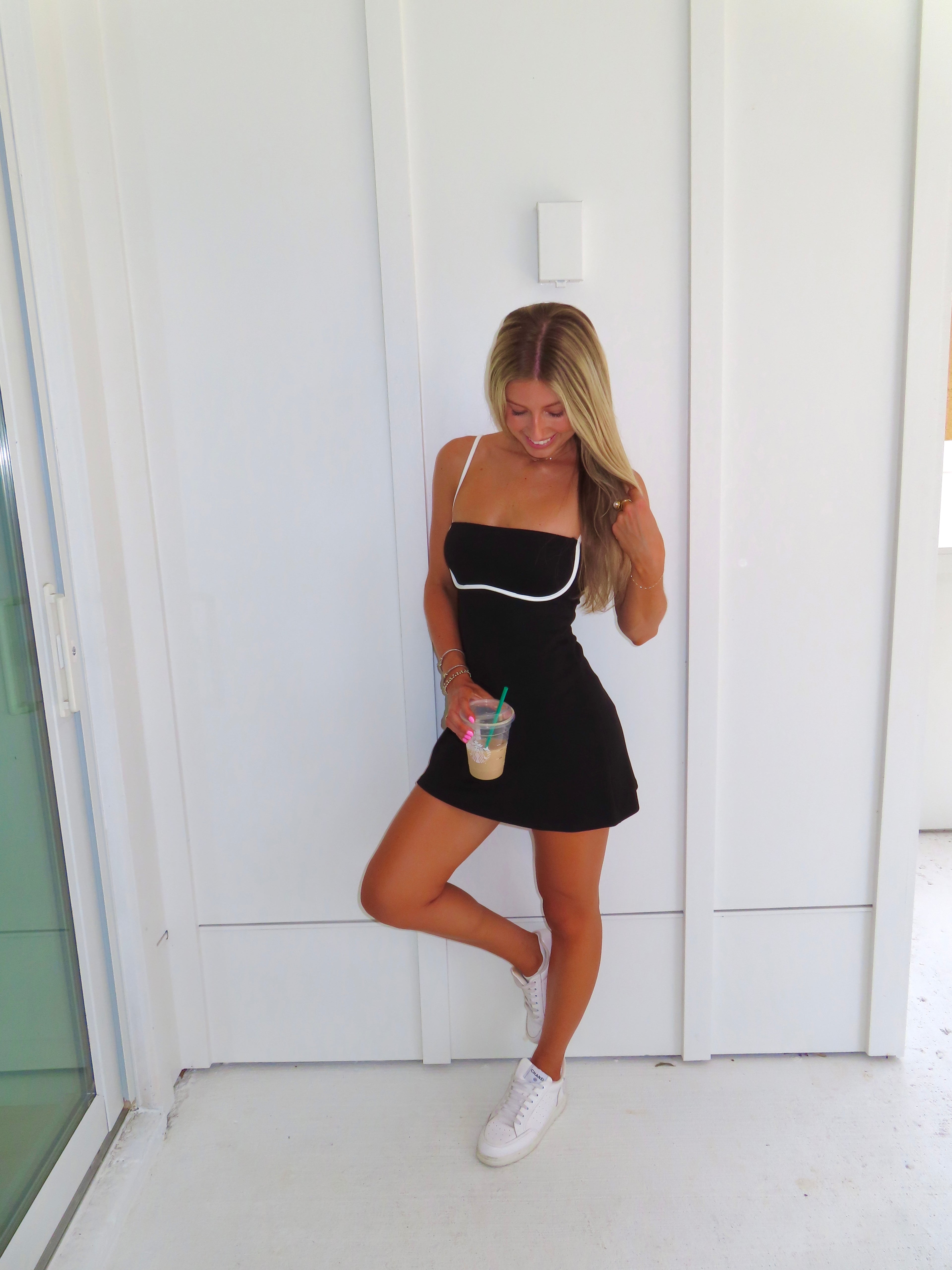 A woman posing in a black athletic dress with white trim, holding a coffee and wearing white sneakers, standing against a white paneled wall.