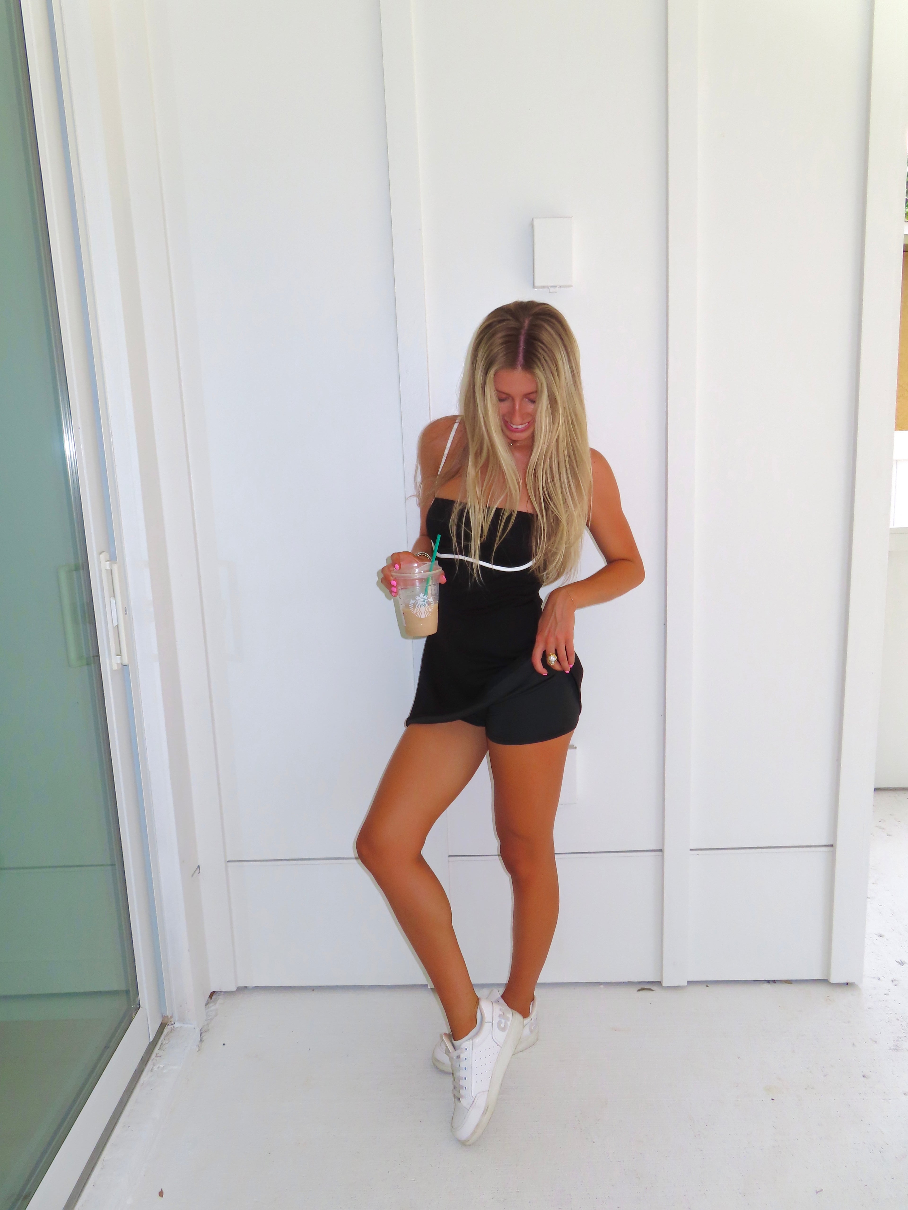 A woman posing in a black athletic dress with white trim and built in shorts, holding a coffee and wearing white sneakers, standing against a white paneled wall.