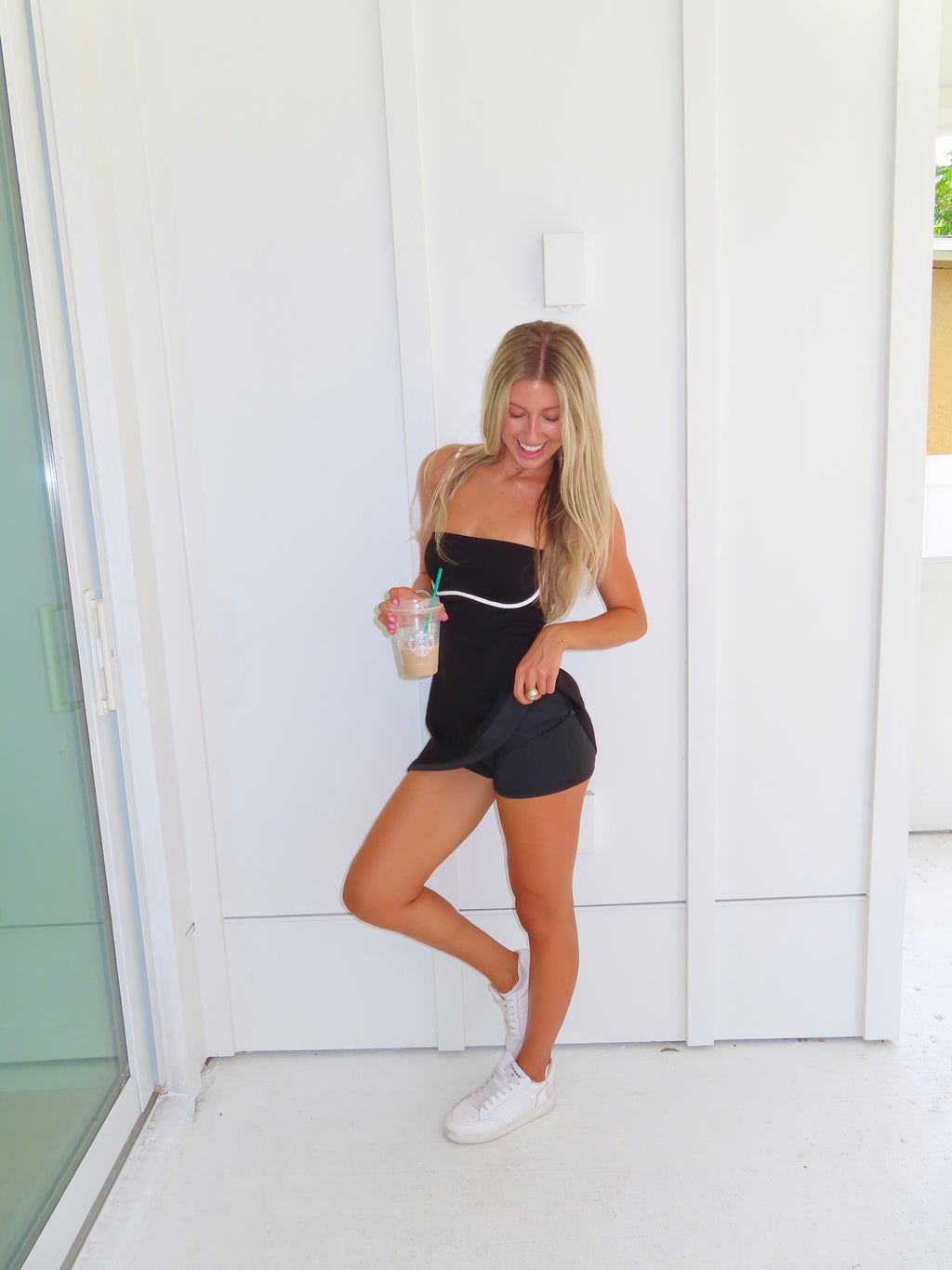 A woman posing in a black athletic dress with white trim and built in shorts, holding a coffee and wearing white sneakers, standing against a white paneled wall.