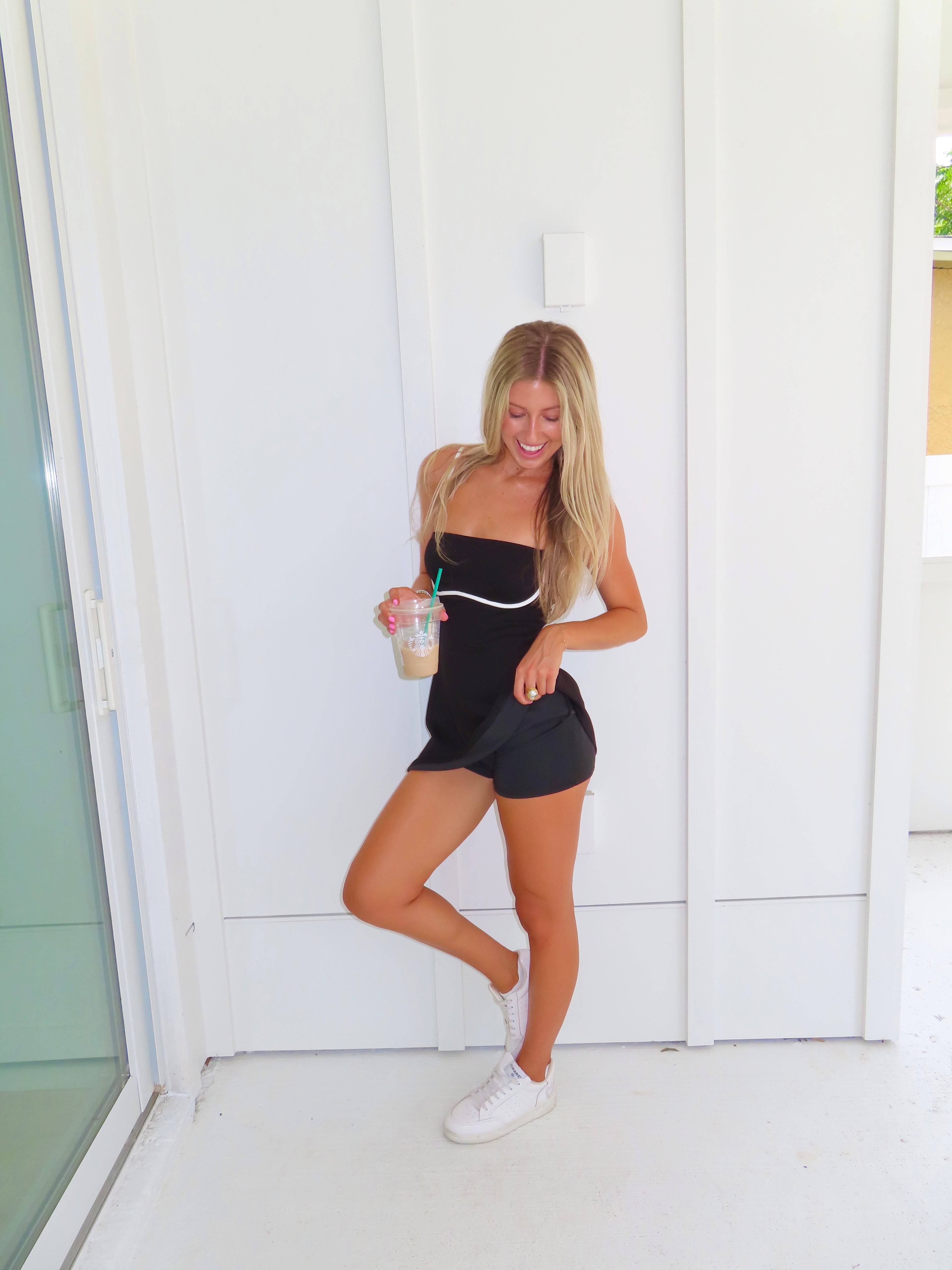 A woman posing in a black athletic dress with white trim and built in shorts, holding a coffee and wearing white sneakers, standing against a white paneled wall.