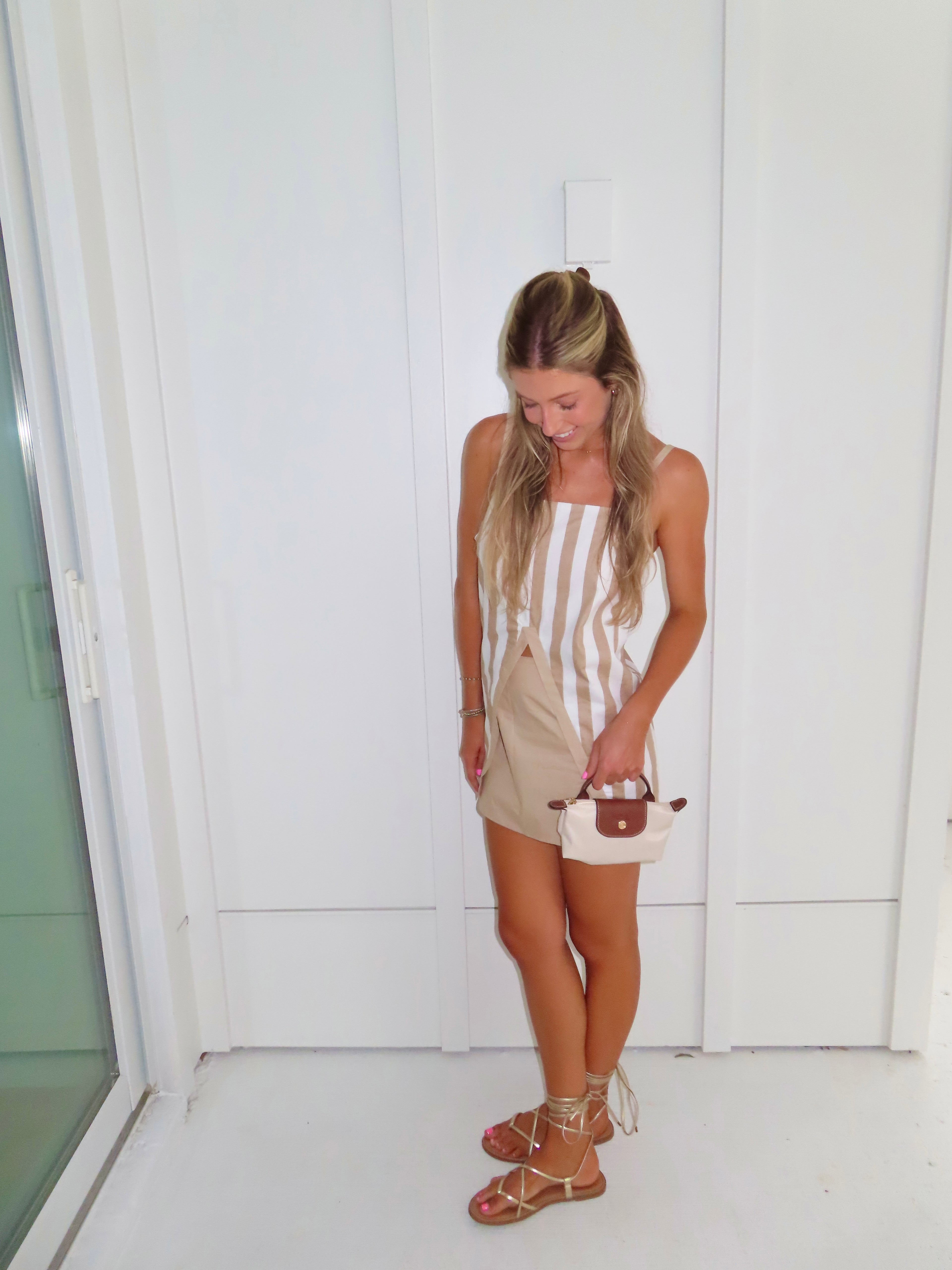 A woman posing in a beige and white vertically striped top with thin spaghetti straps and a tan skort paired with gold sandals and holding a small beige handbag, standing against a white paneled wall.