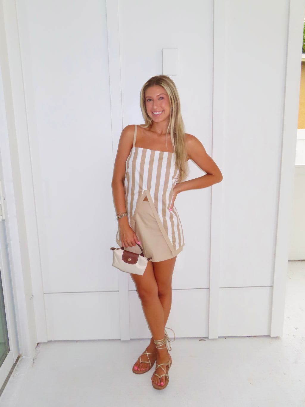 A woman posing in a beige and white vertically striped top with thin spaghetti straps and a tan skort paired with gold sandals and holding a small beige handbag, standing against a white paneled wall.