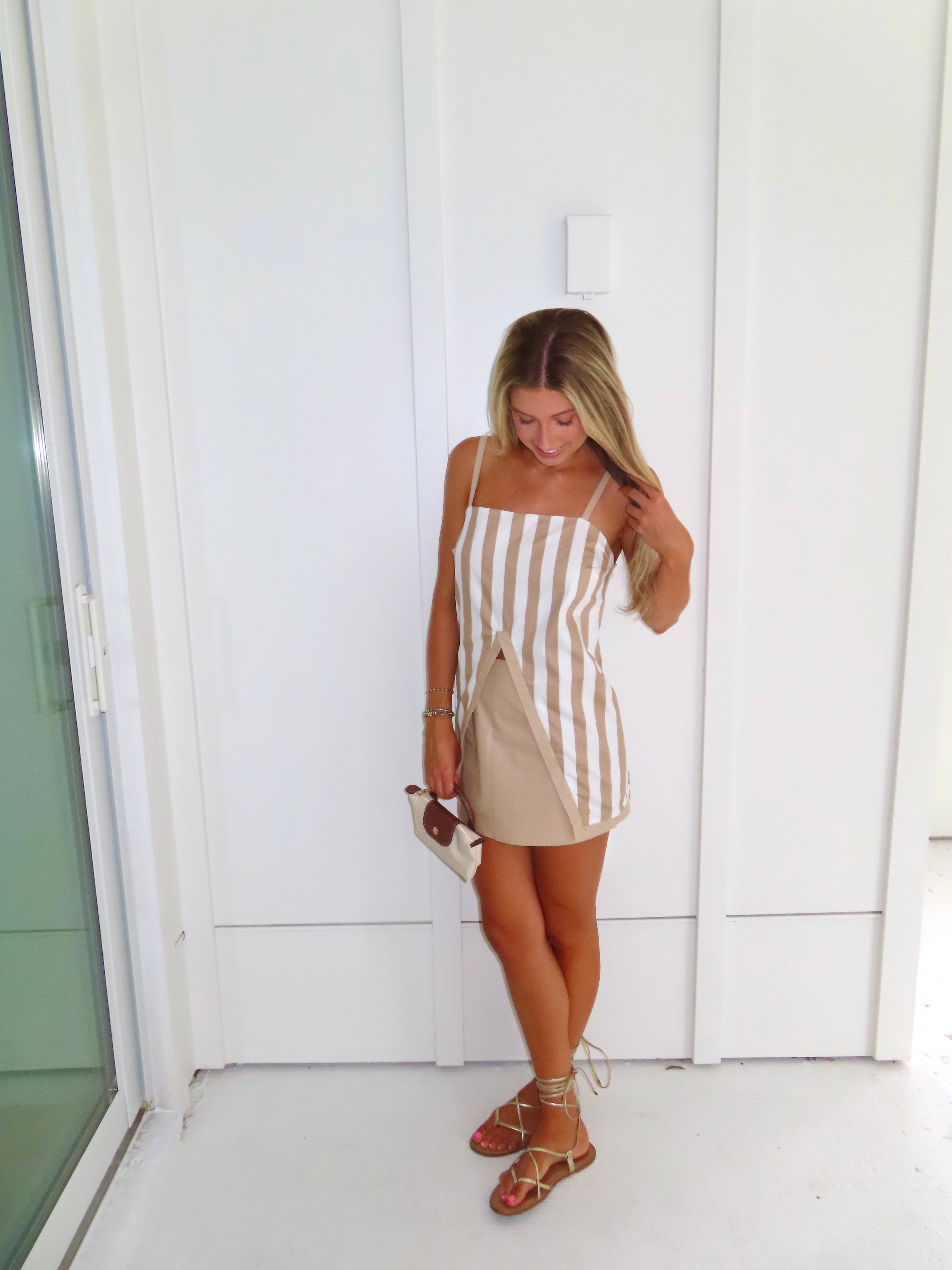 A woman posing in a beige and white vertically striped top with thin spaghetti straps and a tan skort paired with gold sandals and holding a small beige handbag, standing against a white paneled wall.