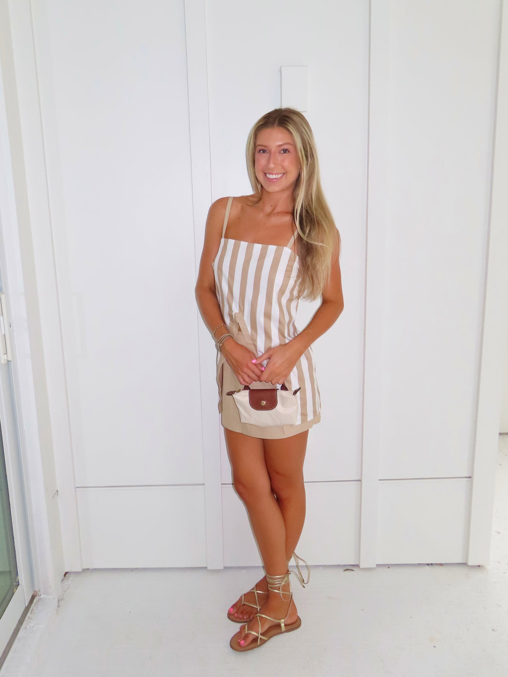 A woman posing in a beige and white vertically striped top with thin spaghetti straps and a tan skort paired with gold sandals and holding a small beige handbag, standing against a white paneled wall.