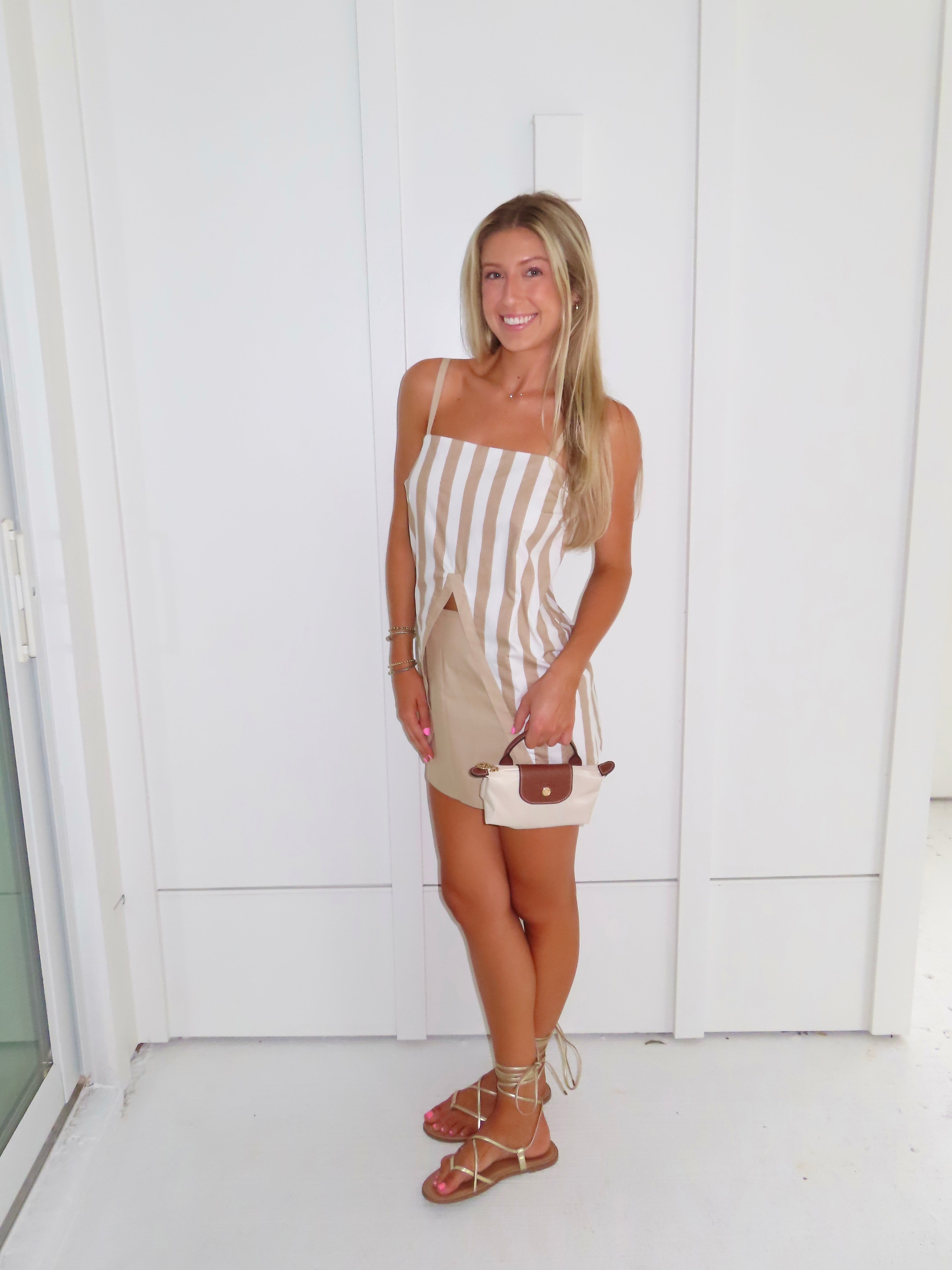 A woman posing in a beige and white vertically striped top with thin spaghetti straps and a tan skort paired with gold sandals and holding a small beige handbag, standing against a white paneled wall.