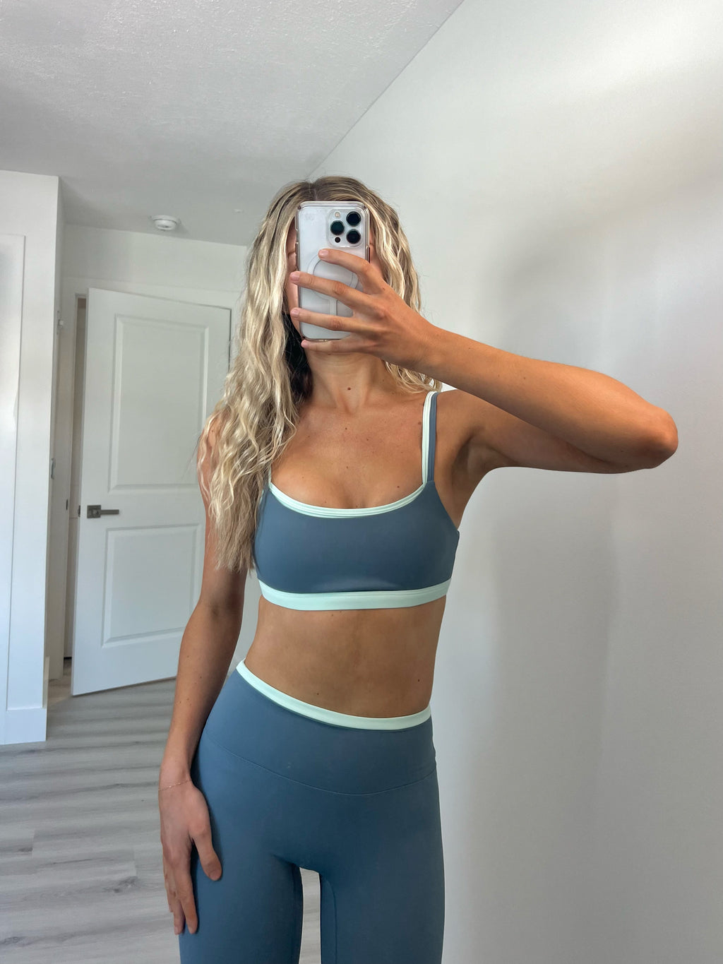 Woman taking a mirror selfie wearing a layered teal and slate blue sports bra and matching high-waisted leggings.