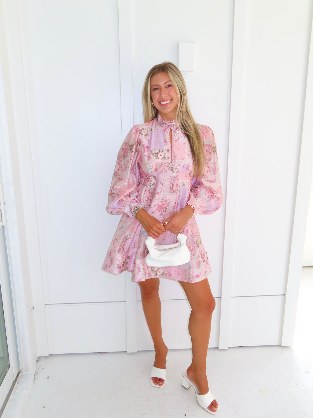 A woman stands outdoors against a white paneled wall wearing a light pink floral mini dress with a high neckline and long balloon sleeves with white heels. She is holding a white purse.