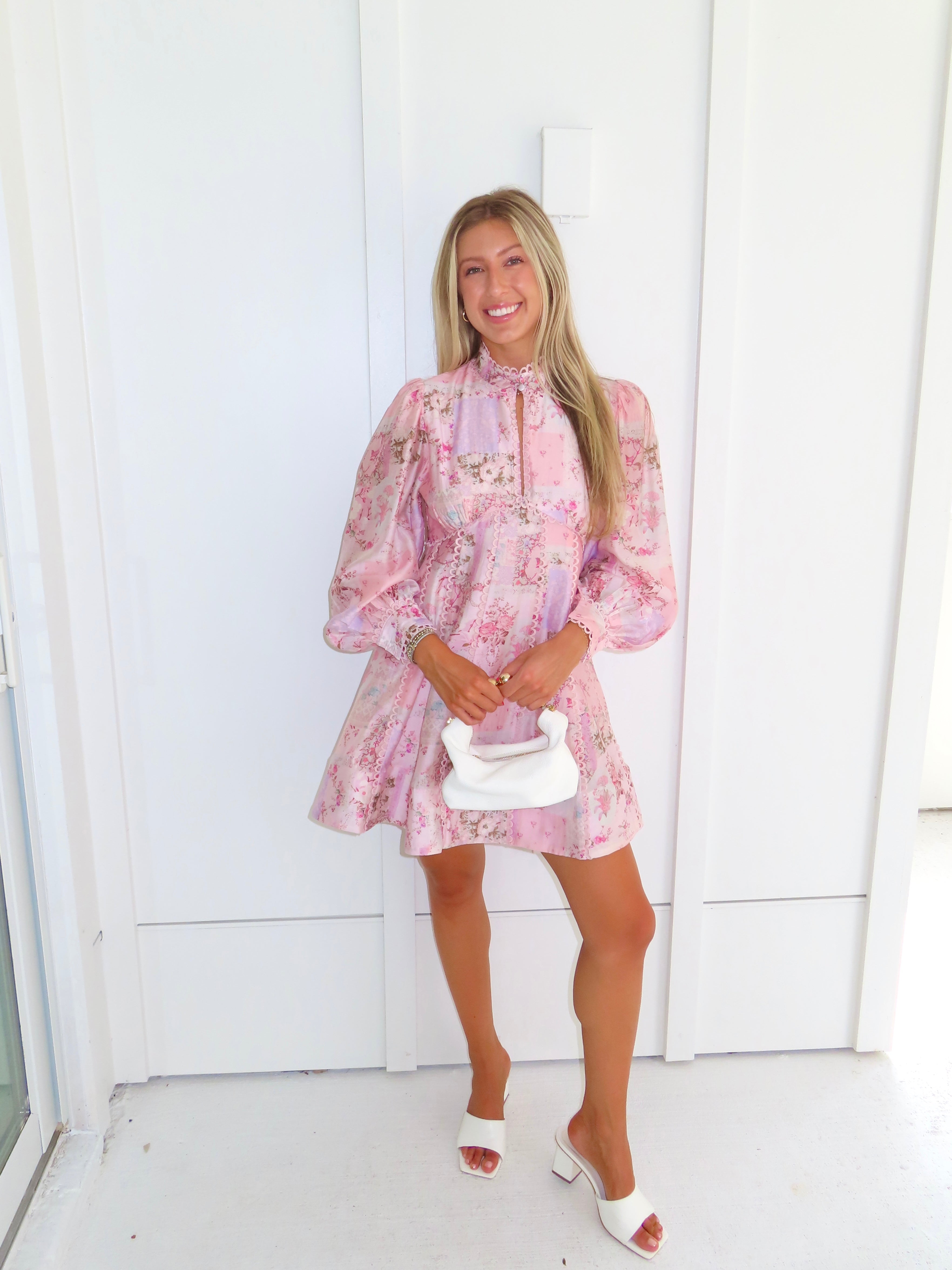 A woman stands outdoors against a white paneled wall wearing a light pink floral mini dress with a high neckline and long balloon sleeves with white heels. She is holding a white purse.