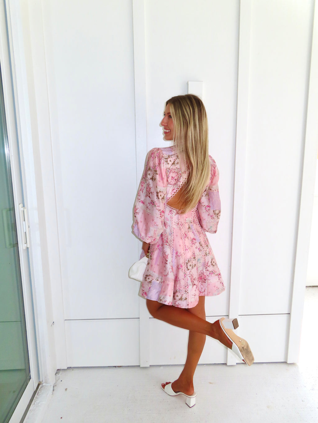 A woman stands outdoors against a white paneled wall wearing a light pink floral mini dress with an open back and long balloon sleeves with white heels. She is holding a white purse.