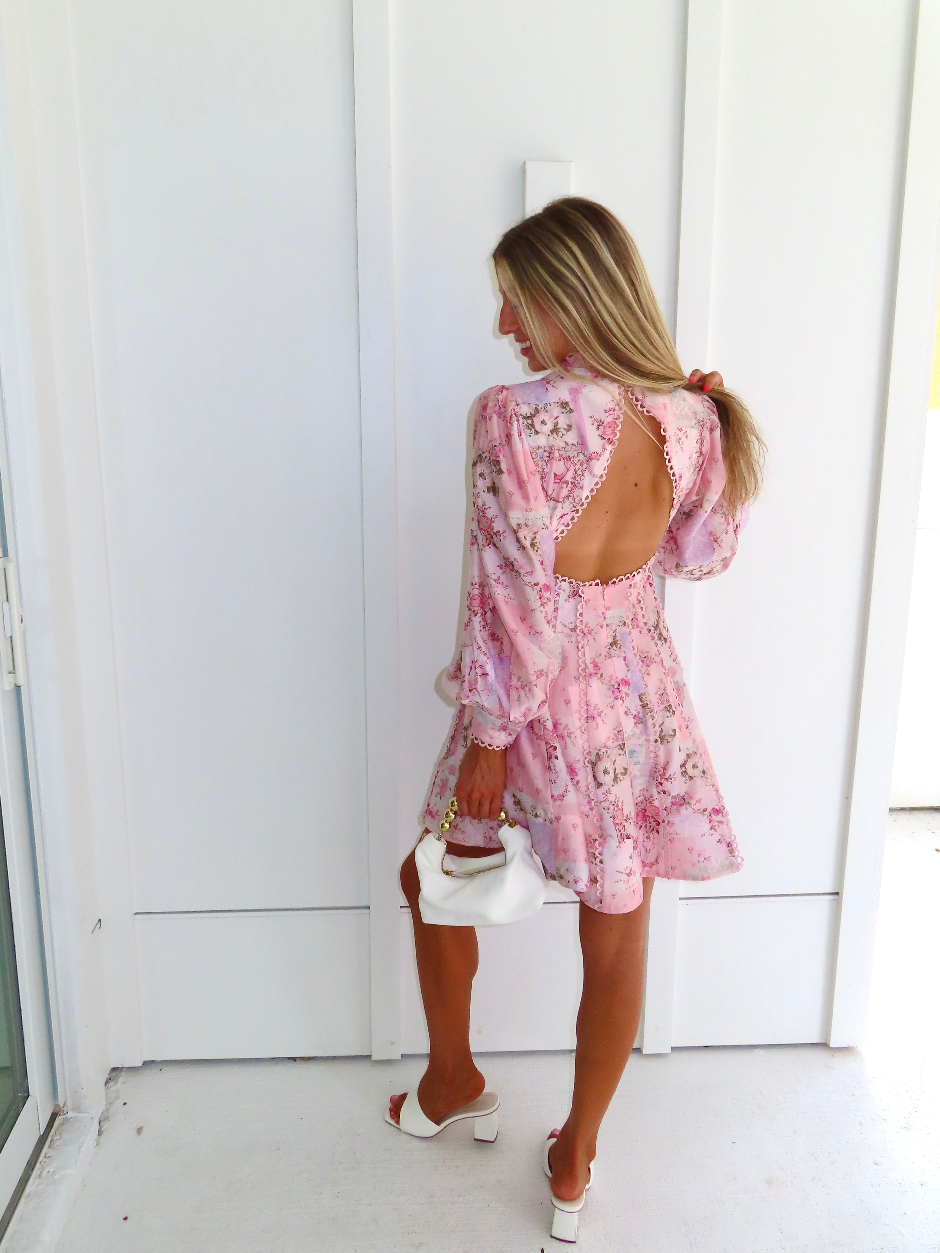 A woman stands outdoors against a white paneled wall wearing a light pink floral mini dress with an open back and long balloon sleeves with white heels. She is holding a white purse.