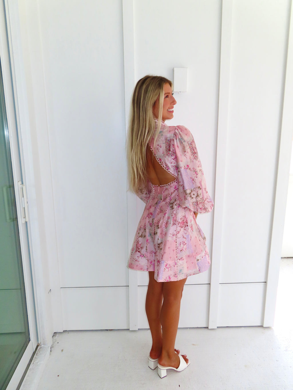 A woman stands outdoors against a white paneled wall wearing a light pink floral mini dress with an open back and long balloon sleeves with white heels. She is holding a white purse.
