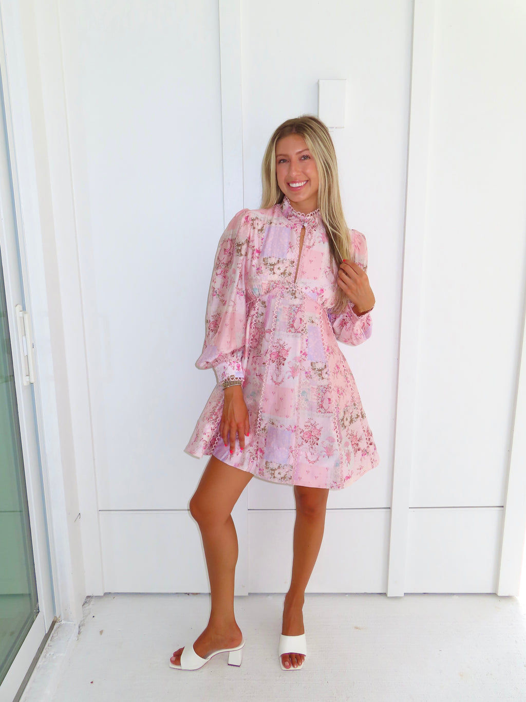 A woman stands outdoors against a white paneled wall wearing a light pink floral mini dress with a high neckline and long balloon sleeves with white heels. 