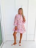 A woman stands outdoors against a white paneled wall wearing a light pink floral mini dress with a high neckline and long balloon sleeves with white heels. 