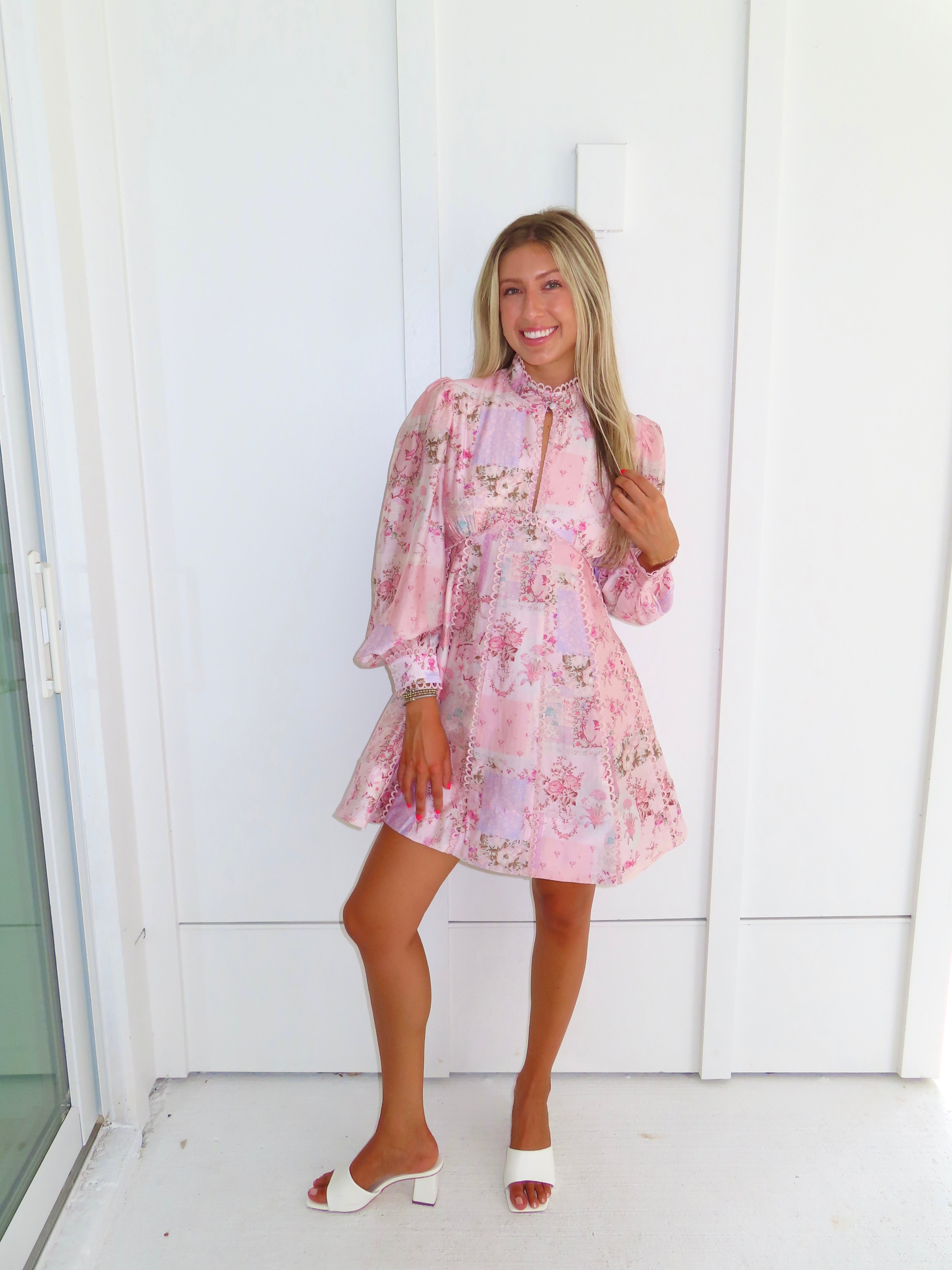 A woman stands outdoors against a white paneled wall wearing a light pink floral mini dress with a high neckline and long balloon sleeves with white heels. 