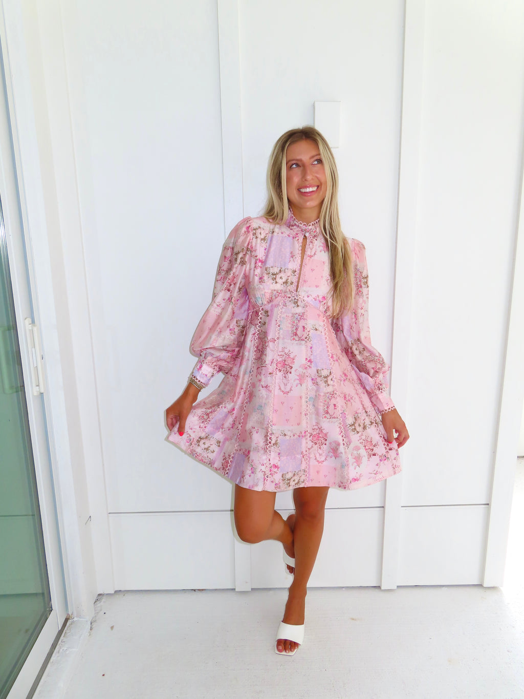 A woman stands outdoors against a white paneled wall wearing a light pink floral mini dress with a high neckline and long balloon sleeves with white heels. 