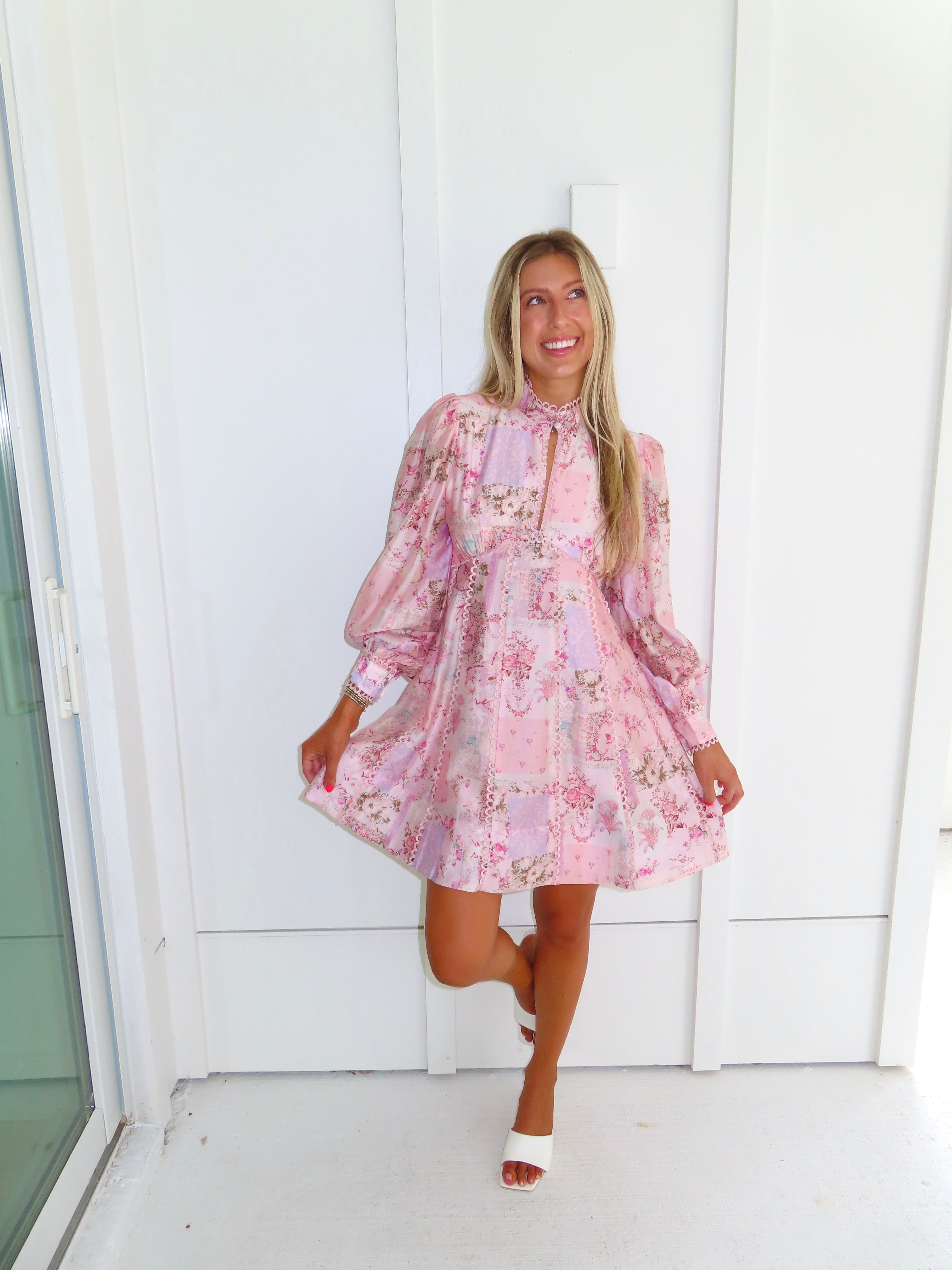 A woman stands outdoors against a white paneled wall wearing a light pink floral mini dress with a high neckline and long balloon sleeves with white heels. 