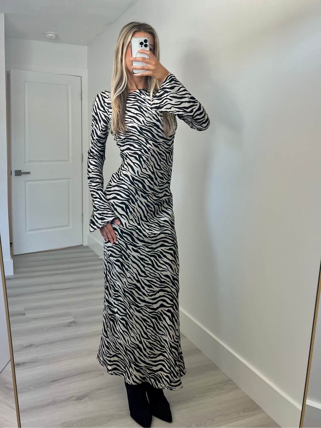 A woman takes a mirror selfie in a room wearing a long-sleeve black and white zebra-print maxi dress paired with black ankle boots.