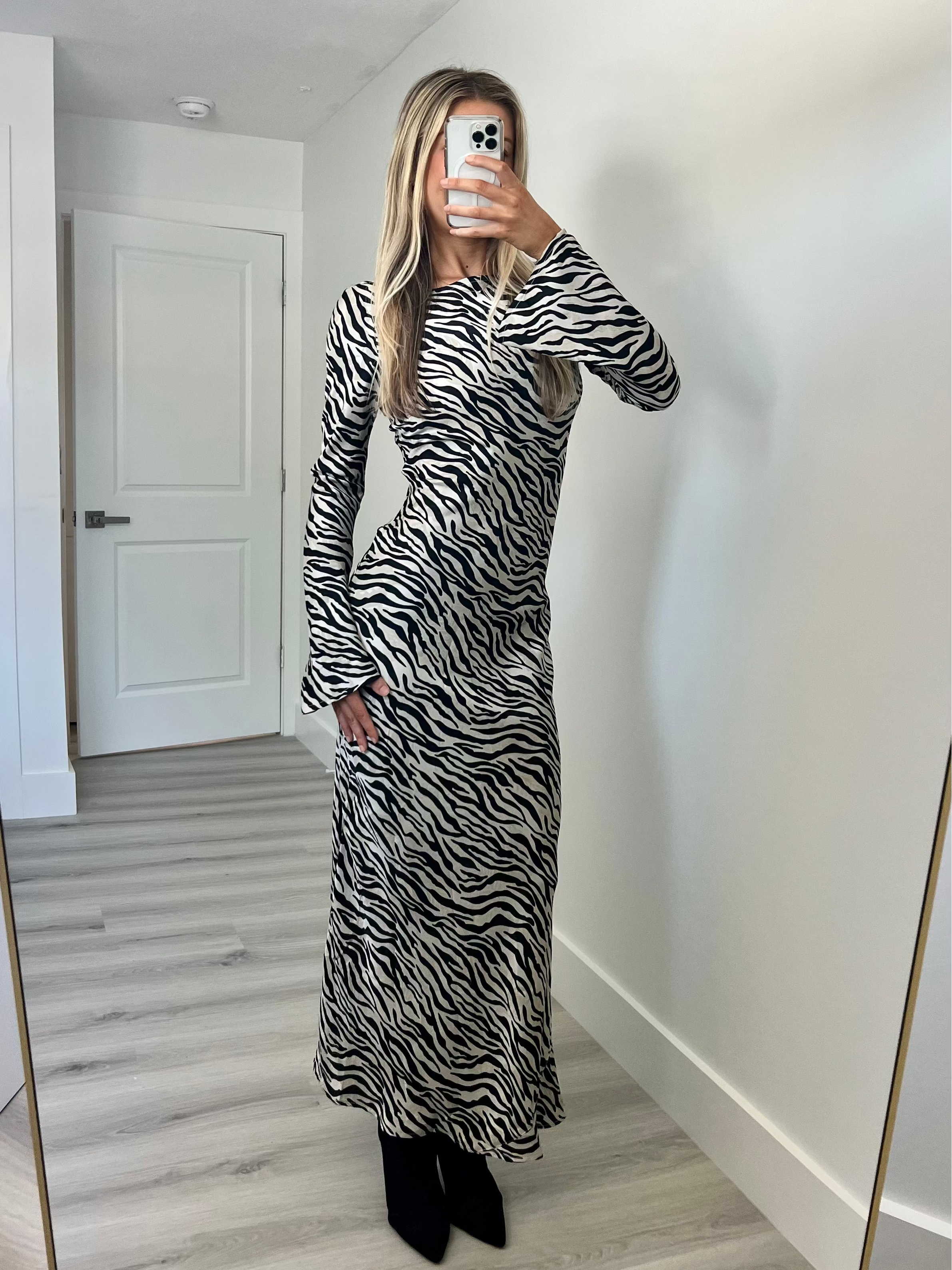A woman takes a mirror selfie in a room wearing a long-sleeve black and white zebra-print maxi dress paired with black ankle boots.