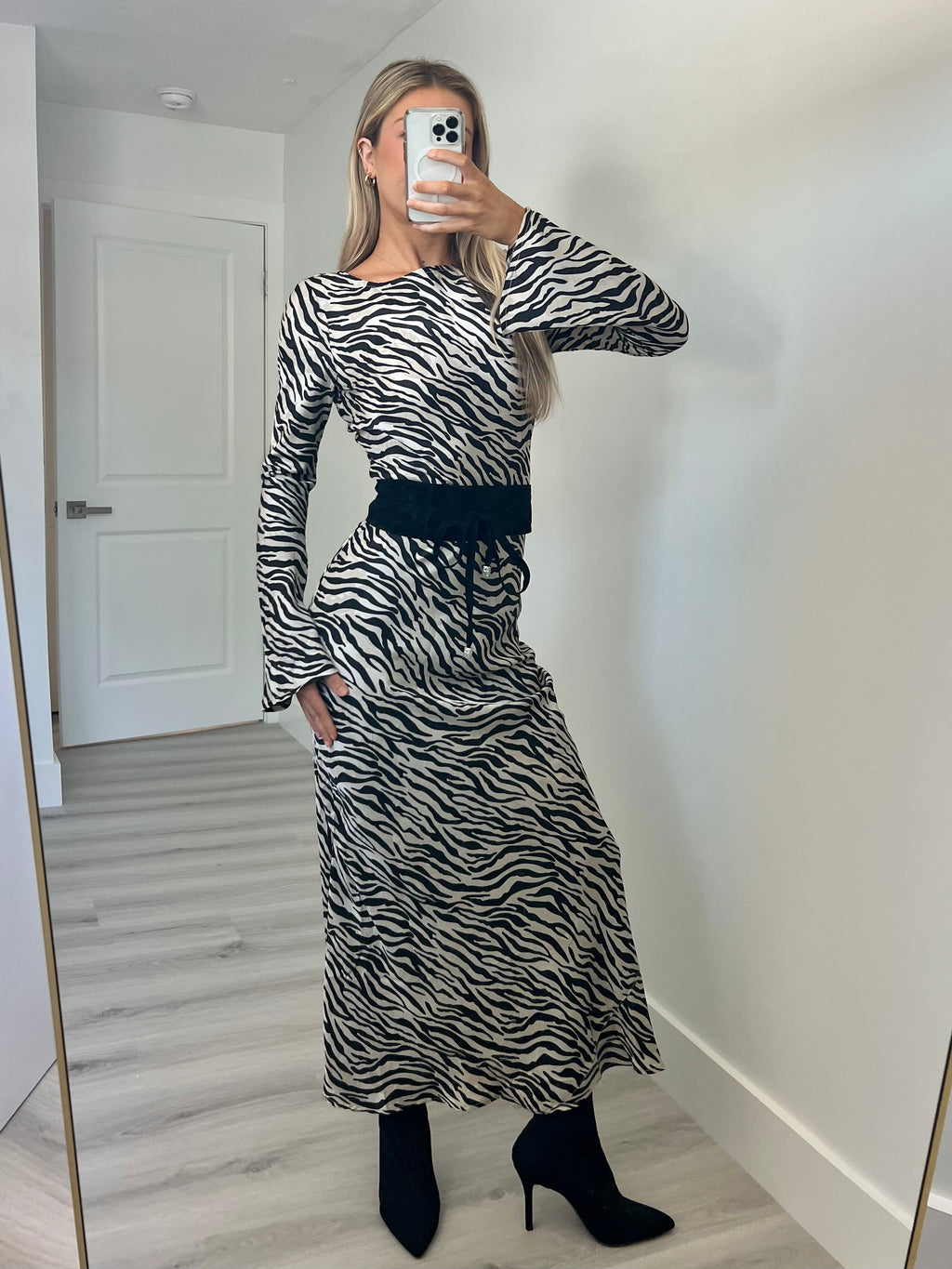A woman takes a mirror selfie in a room wearing a long-sleeve black and white zebra-print maxi dress with a black waist tie, paired with black ankle boots.
