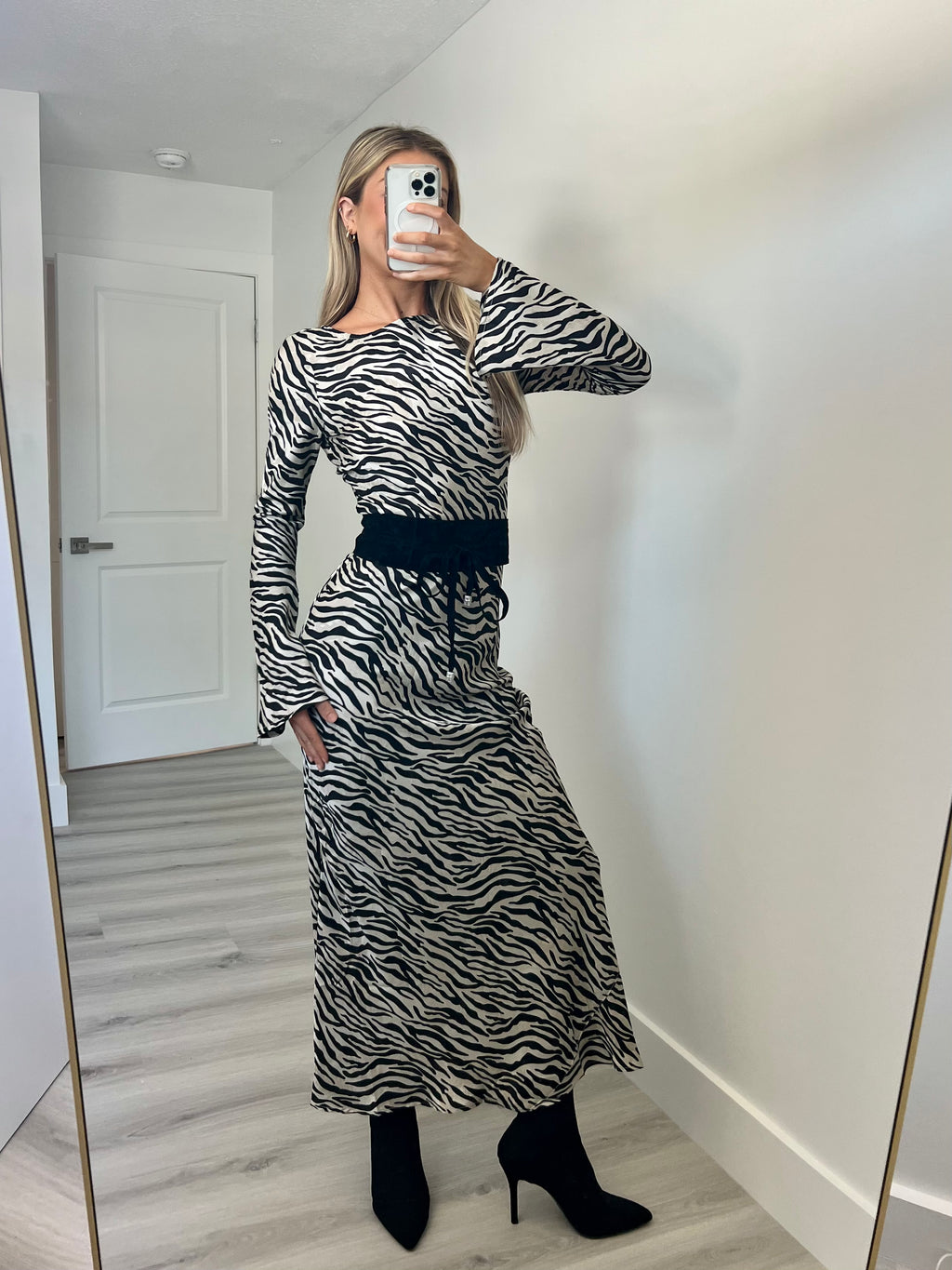 A woman takes a mirror selfie in a room wearing a long-sleeve black and white zebra-print maxi dress with a black waist tie, paired with black ankle boots.