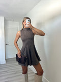A woman takes a mirror selfie wearing a sleeveless, high-neck, flared mini dress in a dark taupe-brown color, paired with beige knee-high boots and holding a dark brown clutch.