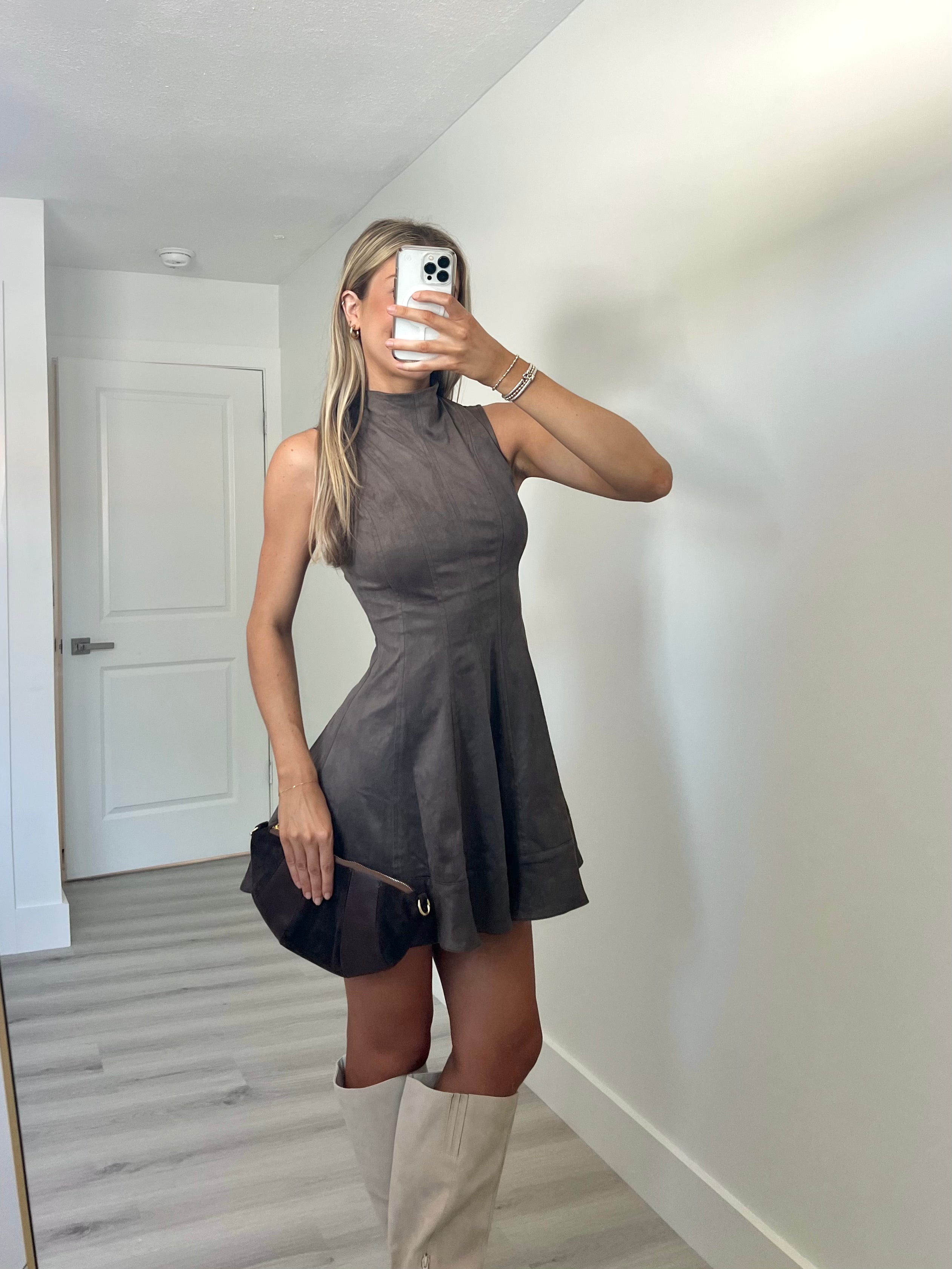 A woman takes a mirror selfie wearing a sleeveless, high-neck, flared mini dress in a dark taupe-brown color, paired with beige knee-high boots and holding a dark brown clutch.