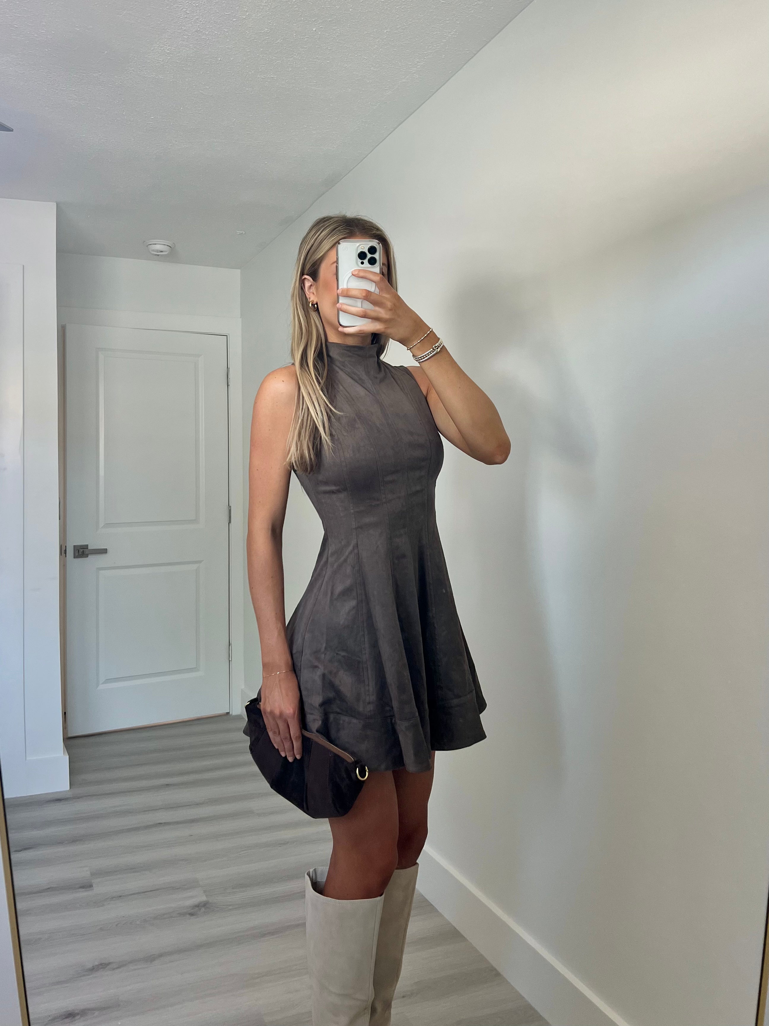 A woman takes a mirror selfie wearing a sleeveless, high-neck, flared mini dress in a dark taupe-brown color, paired with beige knee-high boots and holding a dark brown clutch.
