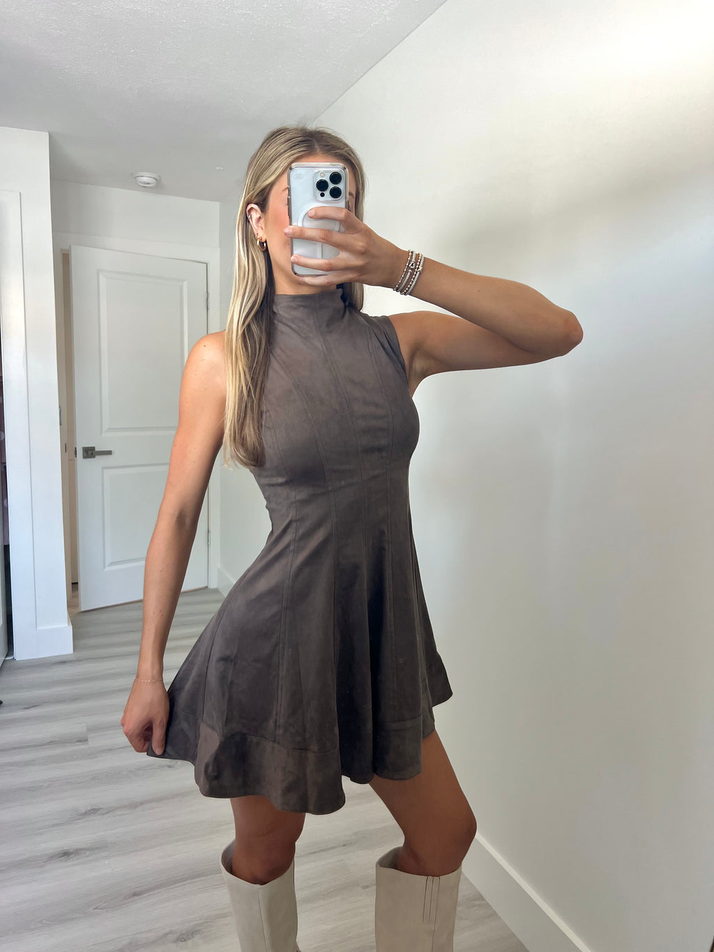 A woman takes a mirror selfie wearing a sleeveless, high-neck, flared mini dress in a dark taupe-brown color, paired with beige knee-high boots.