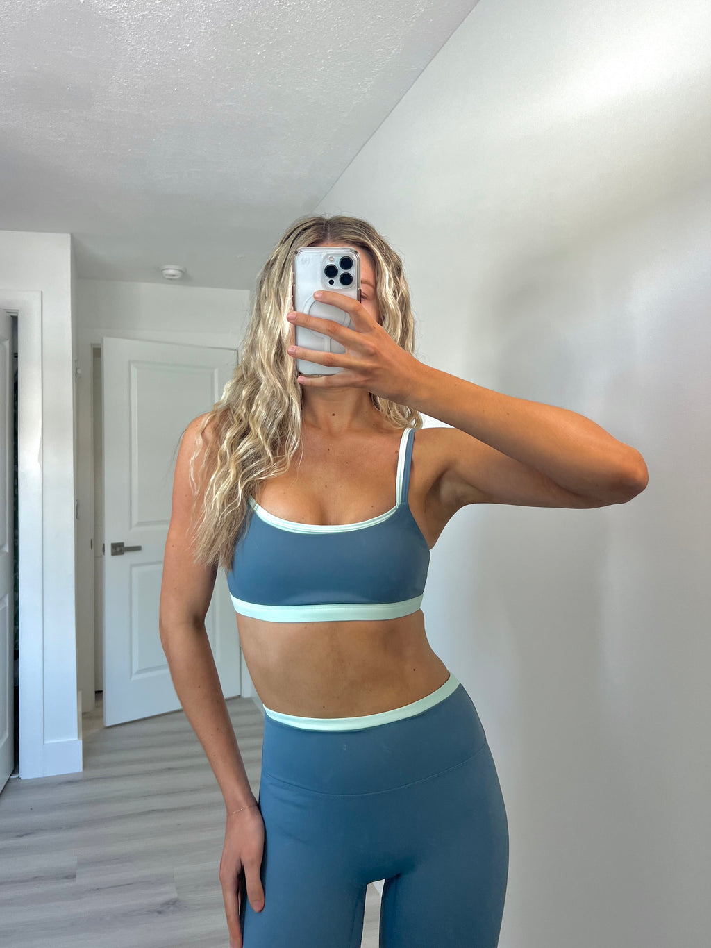 Woman taking a mirror selfie wearing a layered teal and slate blue sports bra and matching high-waisted leggings.