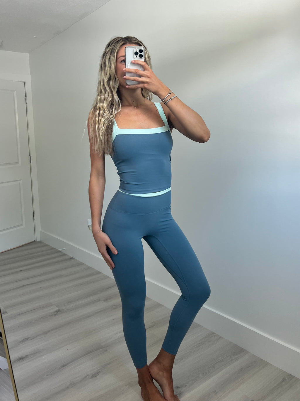 A woman takes a mirror selfie wearing a matching blue activewear set consisting of a scoop-neck tank top and high-waisted leggings with light blue trim, in a room with light walls and wood-look flooring.