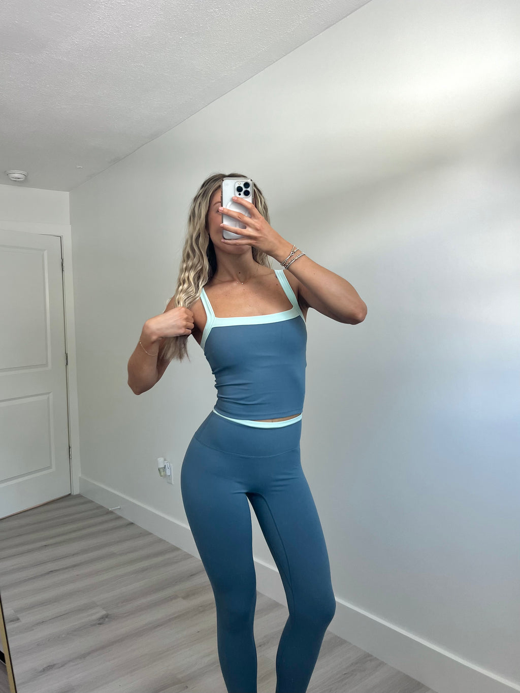 A woman takes a mirror selfie wearing a matching blue activewear set consisting of a scoop-neck tank top and high-waisted leggings with light blue trim, in a room with light walls and wood-look flooring.